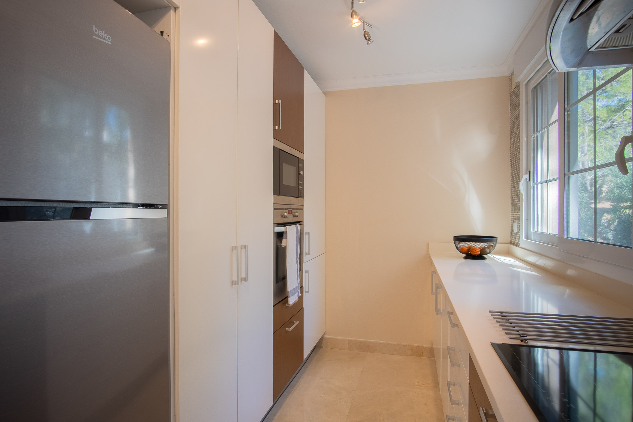 Cuisine moderne avec placards blancs et bois, électroménagers en acier inoxydable intégrés et plan de travail blanc. Une fenêtre latérale offre une lumière naturelle constante.