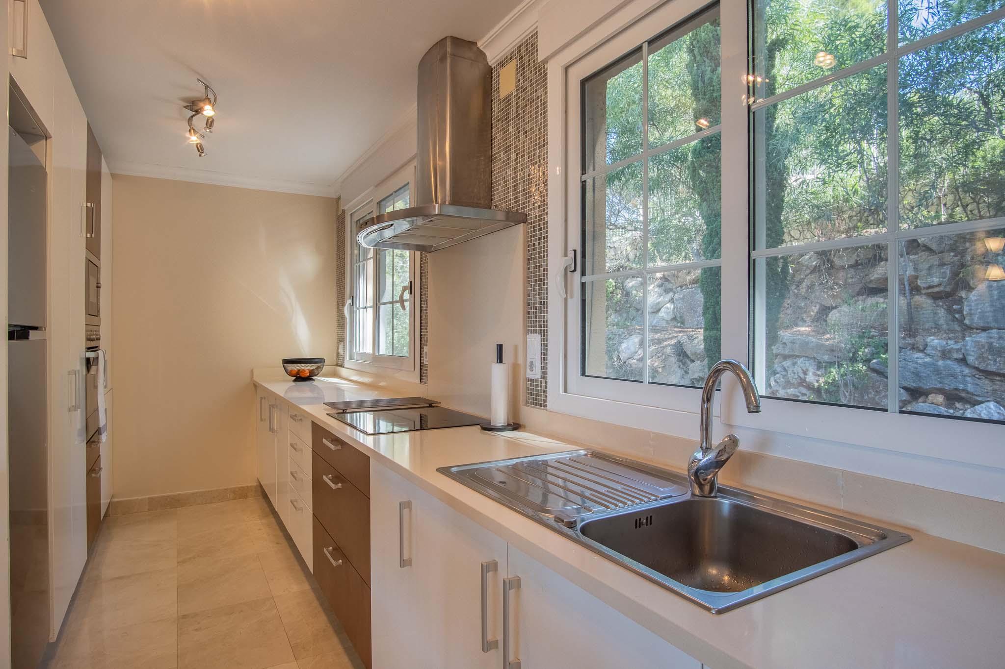 Cuisine moderne avec plan de travail blanc, hotte en inox et grandes fenêtres offrant une vue sur la verdure et les rochers extérieurs.