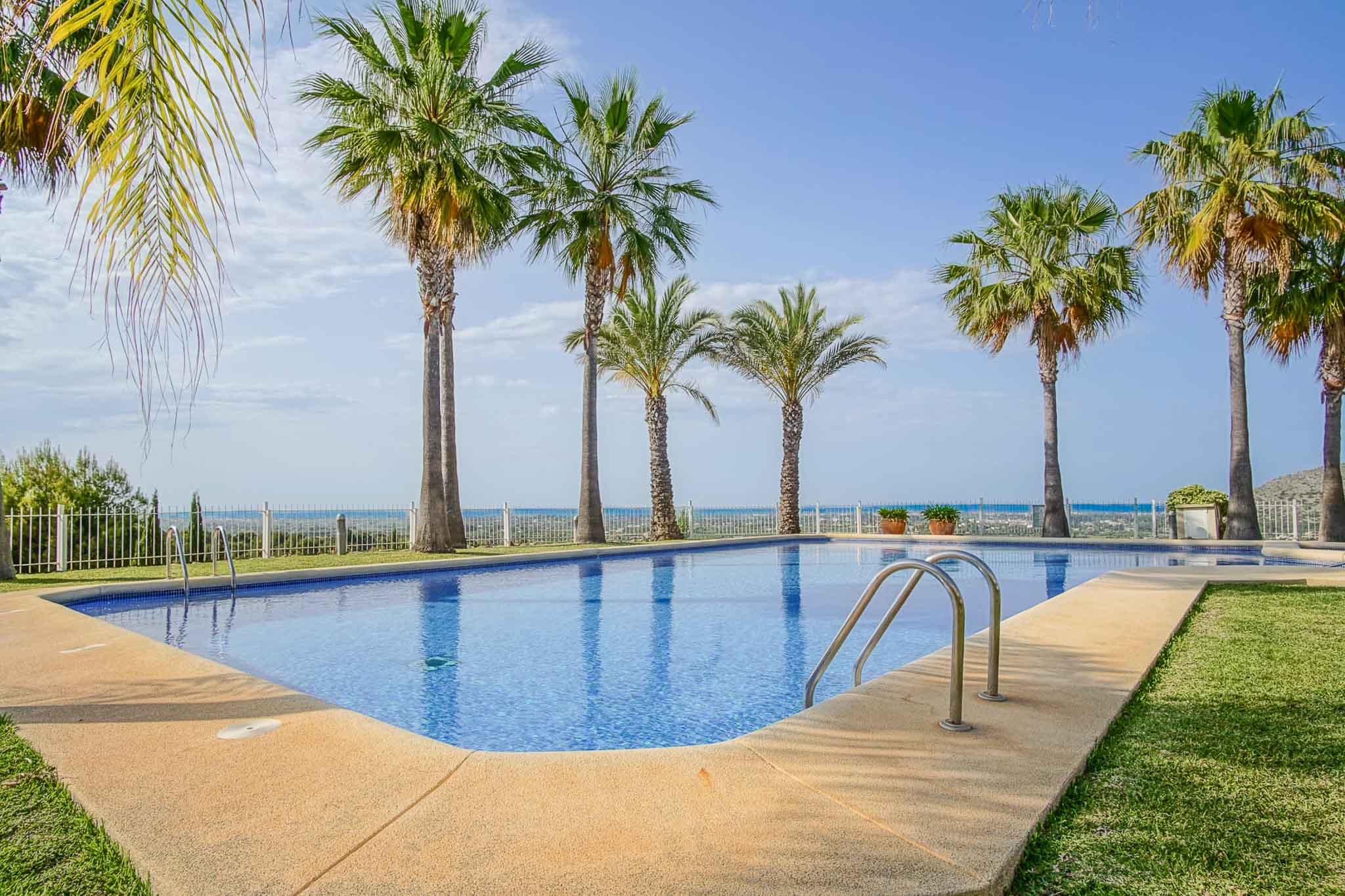 Grande piscine communautaire rectangulaire avec terrasse en pierre, pelouse et palmiers, offrant une vue dégagée sur la côte méditerranéenne et l'horizon.
