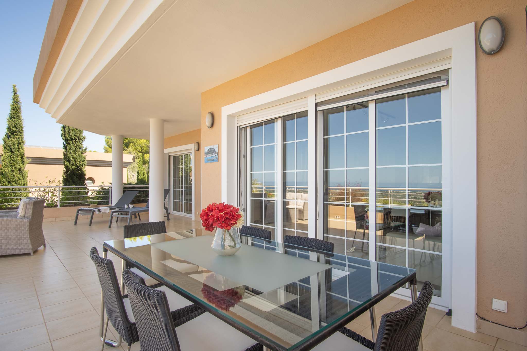 Grande terrasse couverte avec espace repas extérieur, table en verre et chaises en osier, offrant une vue dégagée à travers de larges baies vitrées.