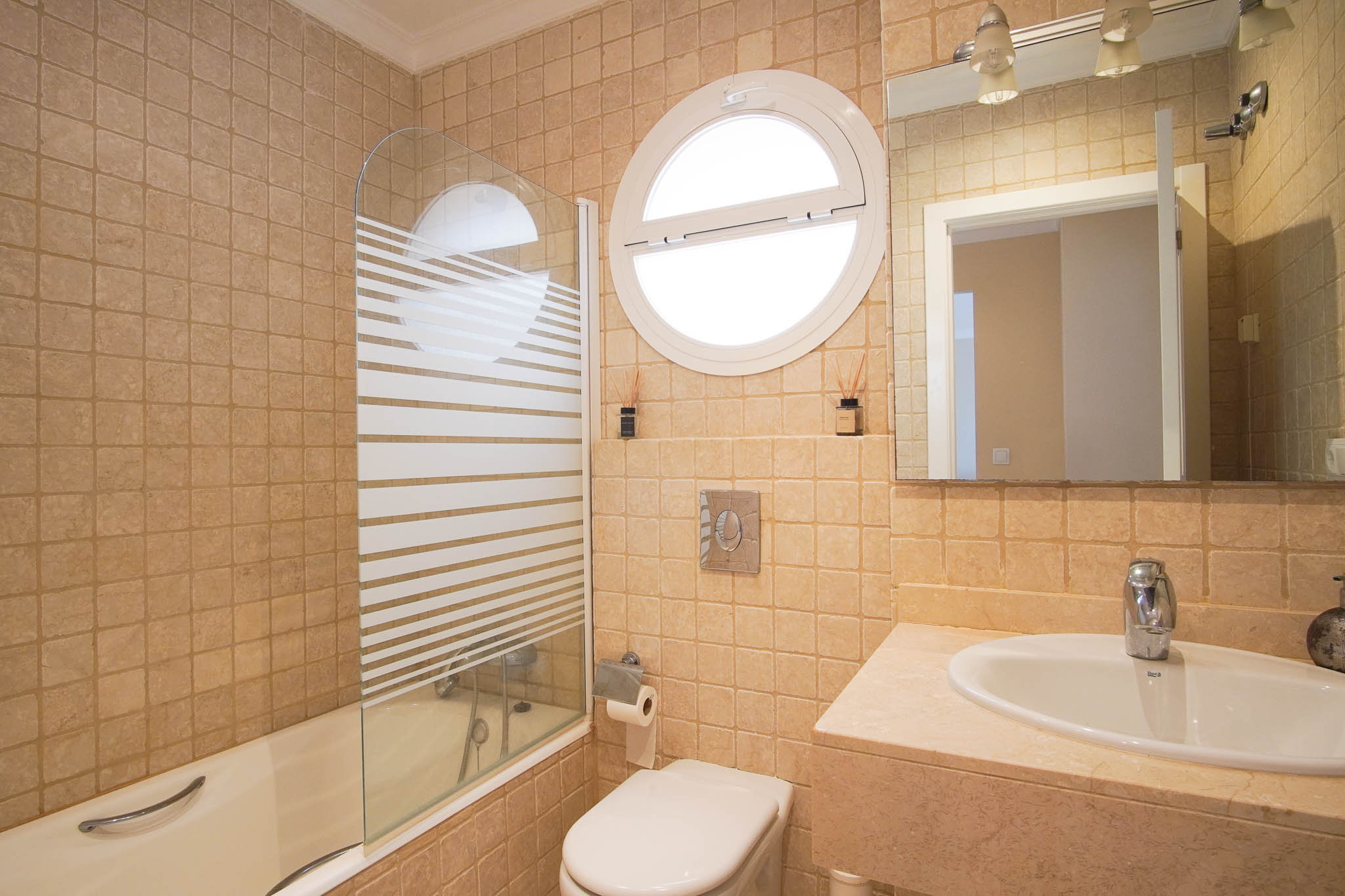 Salle de bain avec finitions en marbre, baignoire avec paroi en verre sérigraphié et une fenêtre circulaire distinctive apportant de la lumière naturelle.