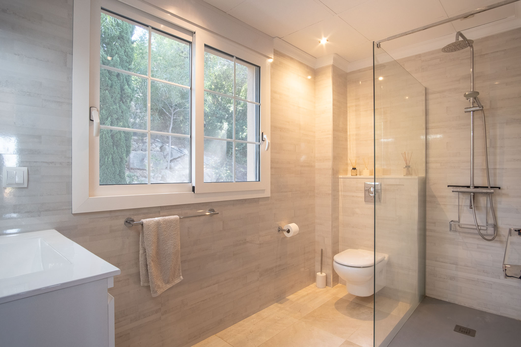 Salle de bain moderne avec douche à l'italienne, paroi en verre et carrelage aux tons neutres. Équipée de sanitaires suspendus et d'une fenêtre offrant une lumière naturelle et vue sur la verdure.