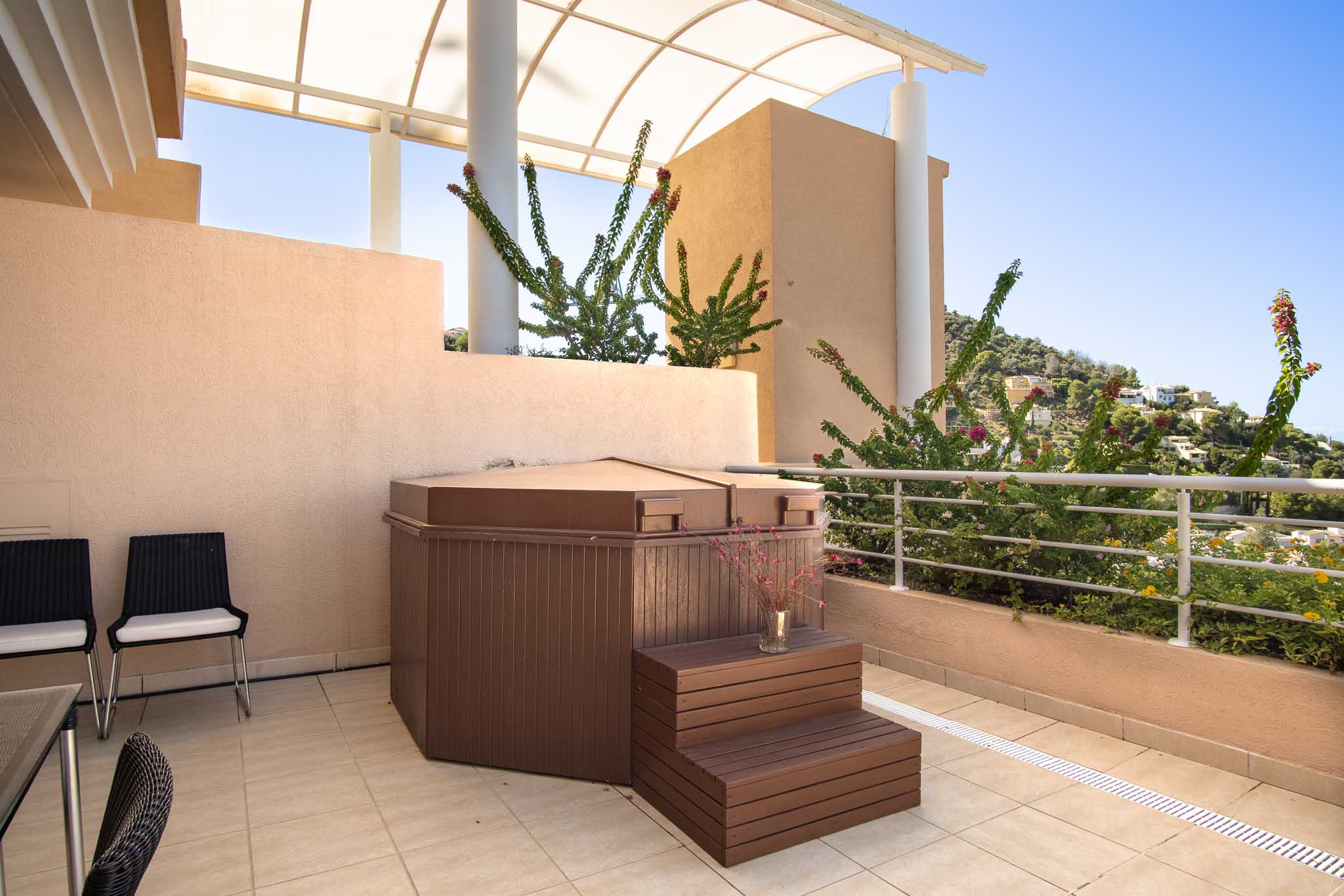 Terrasse privée avec jacuzzi couvert et marches en bois, sous une pergola arquée offrant une vue sur les collines environnantes et la végétation méditerranéenne.