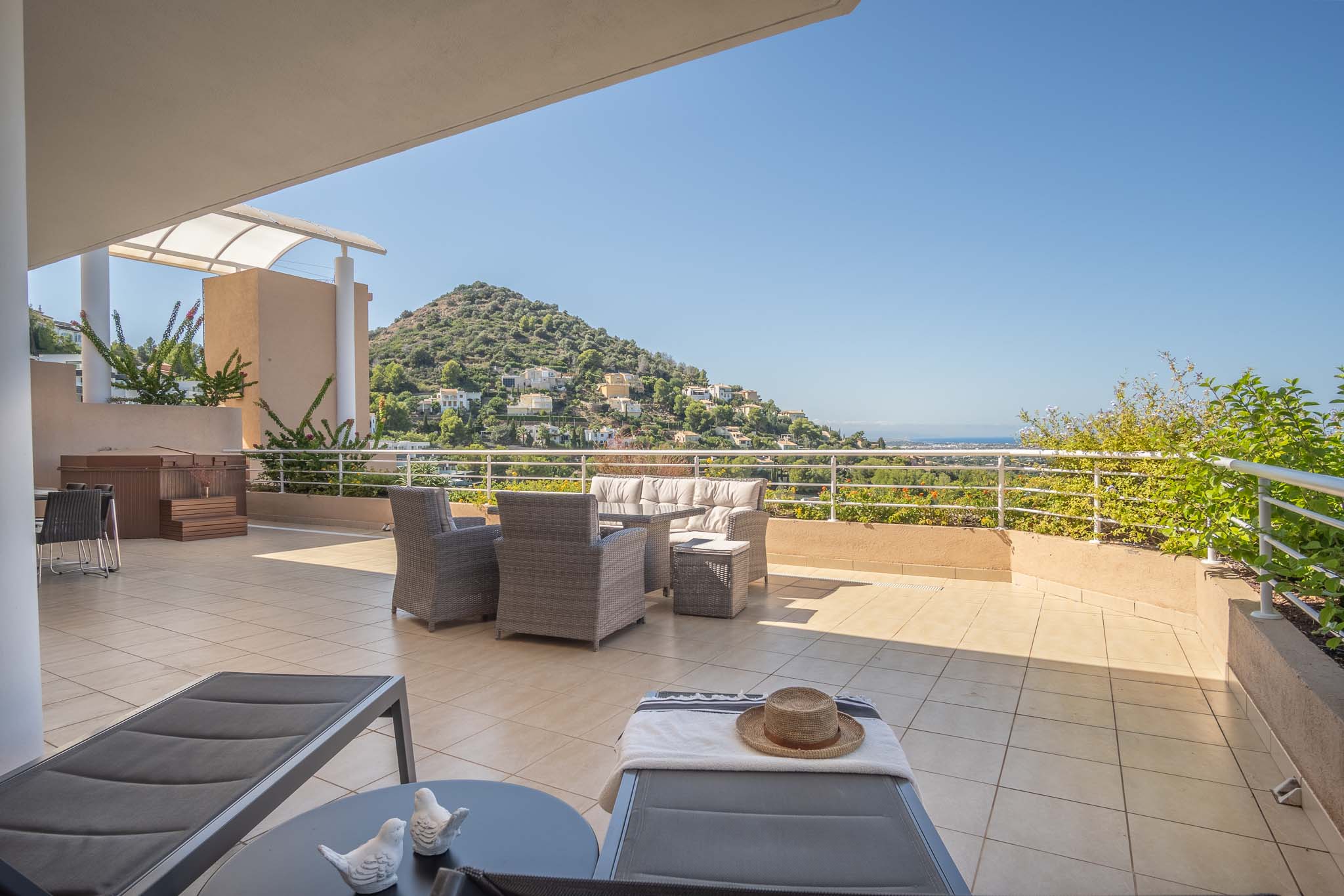 Vaste terrasse carrelée avec salon en résine tressée, jacuzzi privé et bains de soleil. Elle offre une vue panoramique sur les collines et la mer Méditerranée.