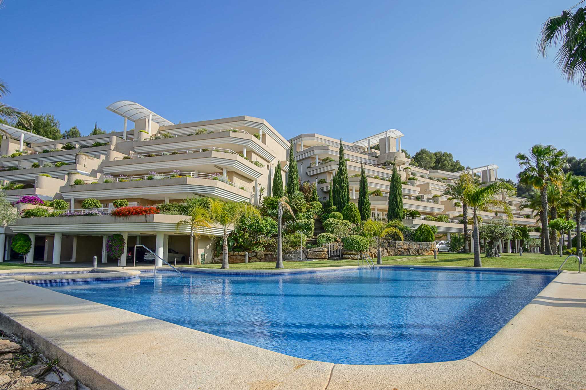 Vue extérieure du complexe Las Mimosas montrant la piscine communautaire et l'architecture en terrasses entourée de jardins méditerranéens et de palmiers.