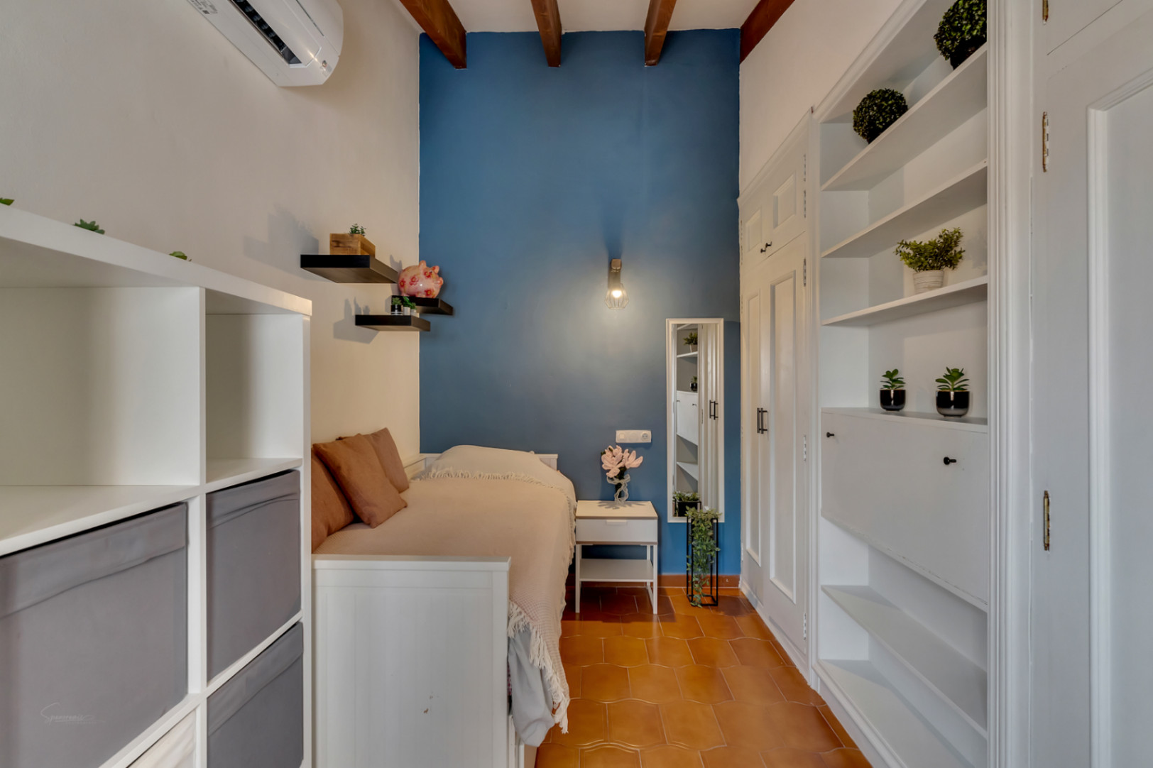 Chambre avec mur d'accent bleu, étagères blanches et sol en terre cuite. Poutres apparentes au plafond.