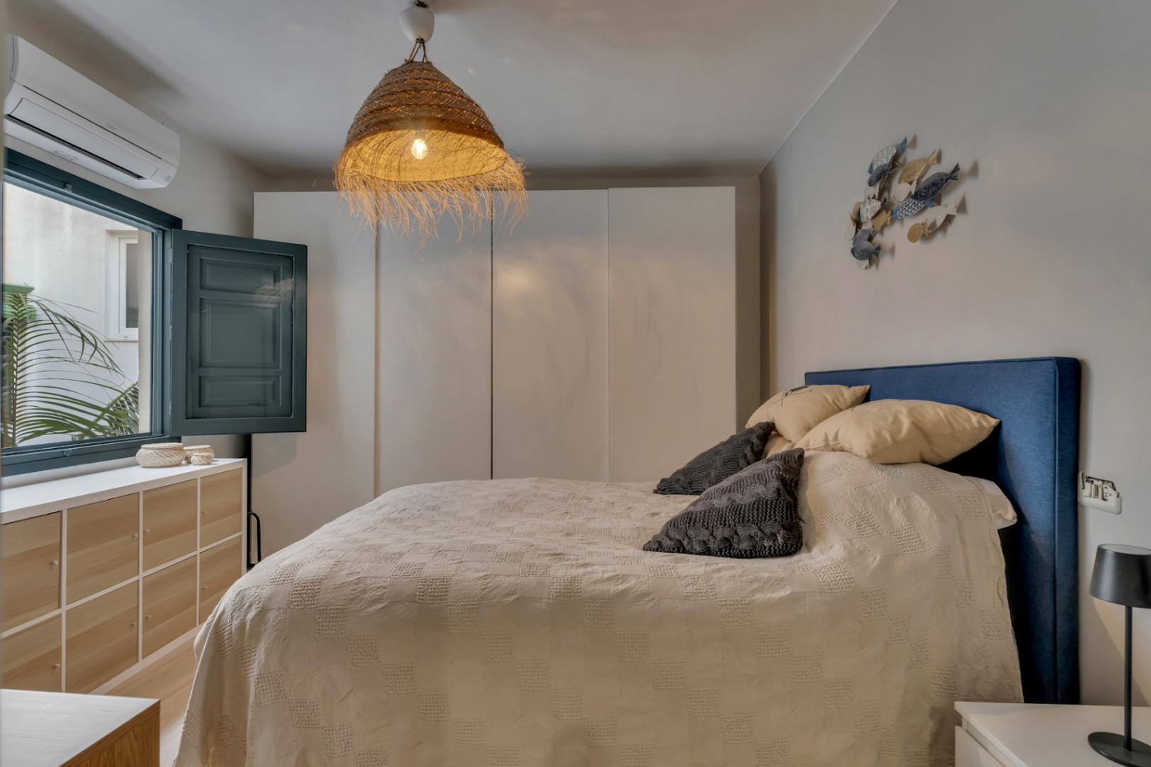 Chambre lumineuse avec tête de lit bleue, lampe en osier et fenêtre à volets.