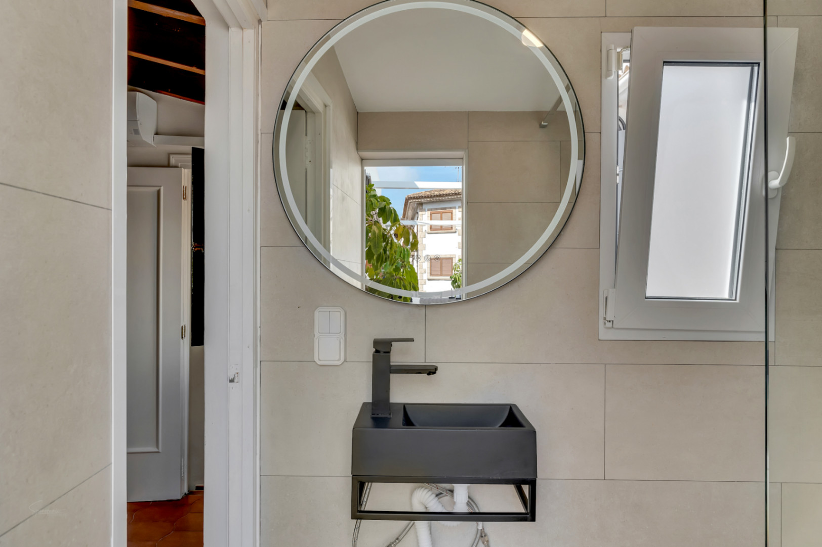 Détail de la salle de bain avec lavabo noir suspendu, miroir rond lumineux et fenêtre à auvent.