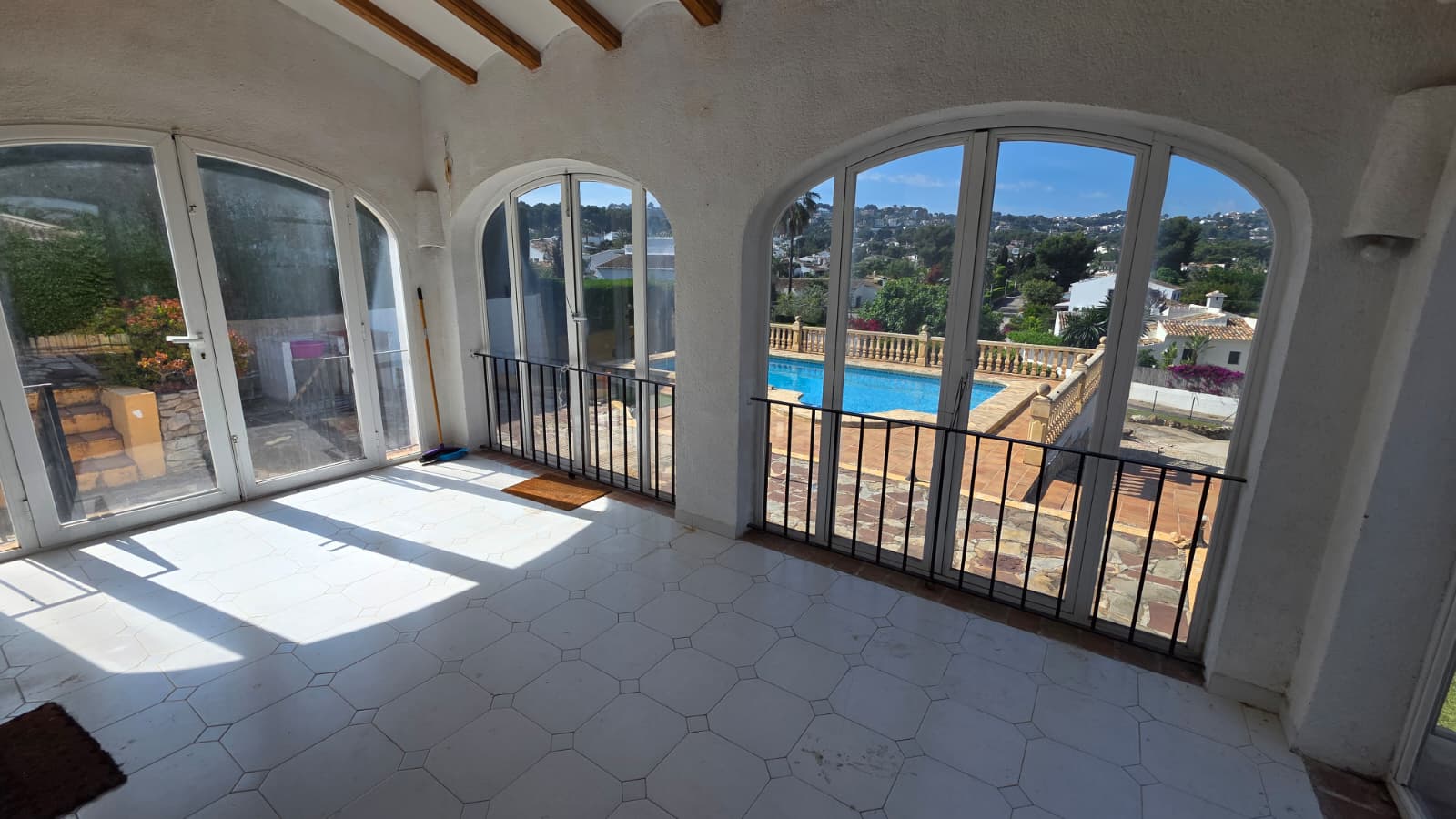 Naya vitrée avec ouvertures en arche, carrelage blanc et poutres apparentes, offrant une vue sur la piscine privée et le quartier résidentiel.