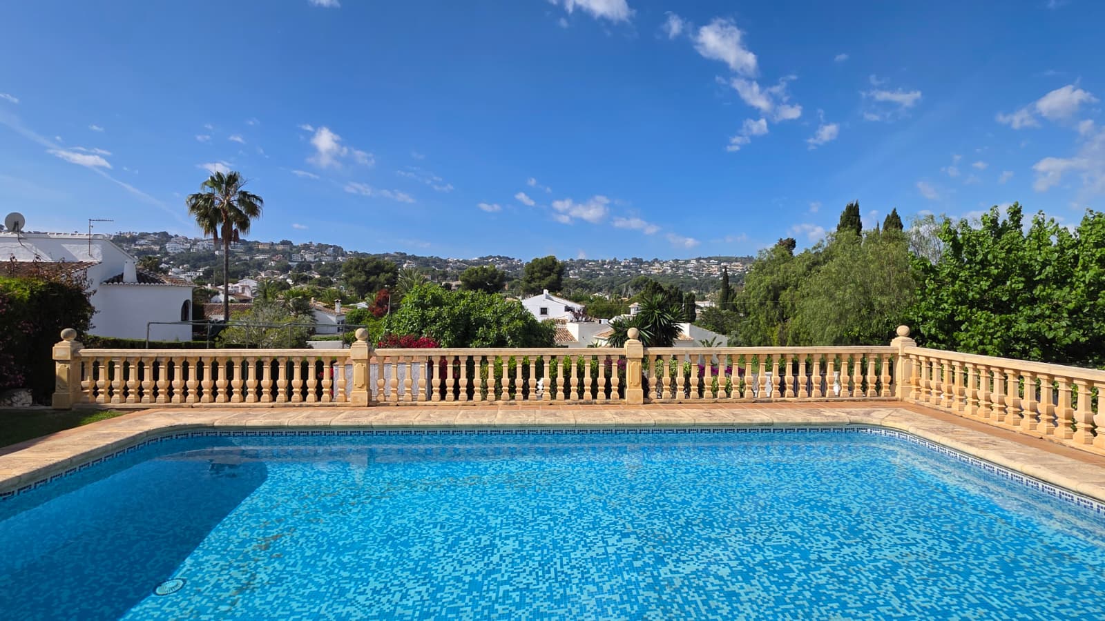 Piscine extérieure avec mosaïque bleue et balustrade en pierre classique, offrant une vue dégagée sur les collines et le quartier résidentiel de Jávea.