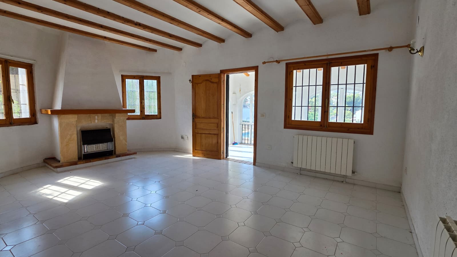 Salon avec poutres apparentes en bois, cheminée en pierre naturelle et sol en carrelage blanc. Accès direct vers l'extérieur par une porte en bois massif.