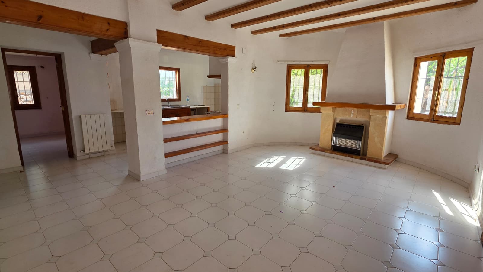 Salon spacieux avec poutres en bois apparentes, cheminée en pierre avec insert et sol en carrelage blanc. Une ouverture type passe-plat relie la pièce à la cuisine.