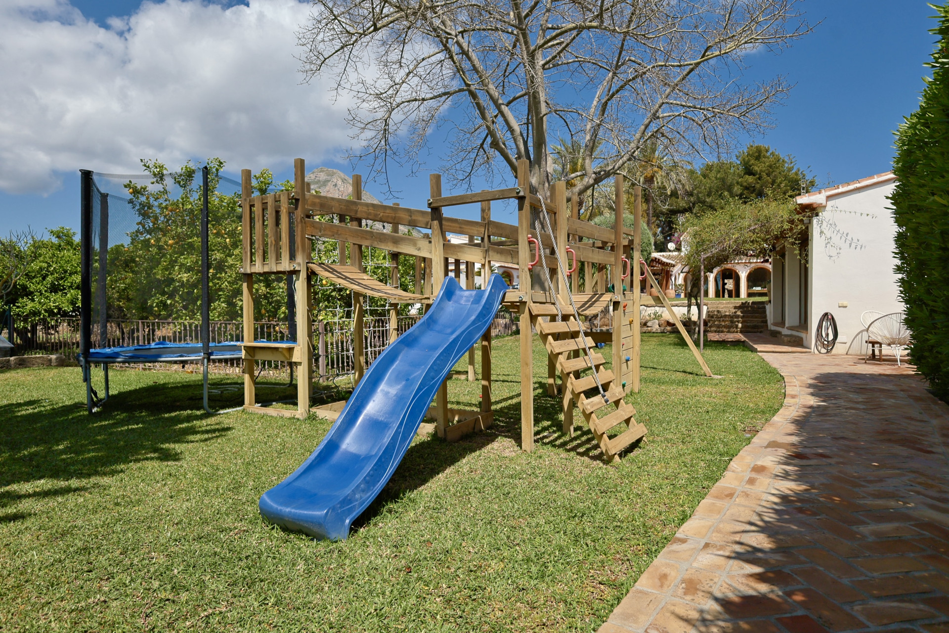 Aire de jeux avec structure en bois, toboggan bleu et trampoline dans le jardin.