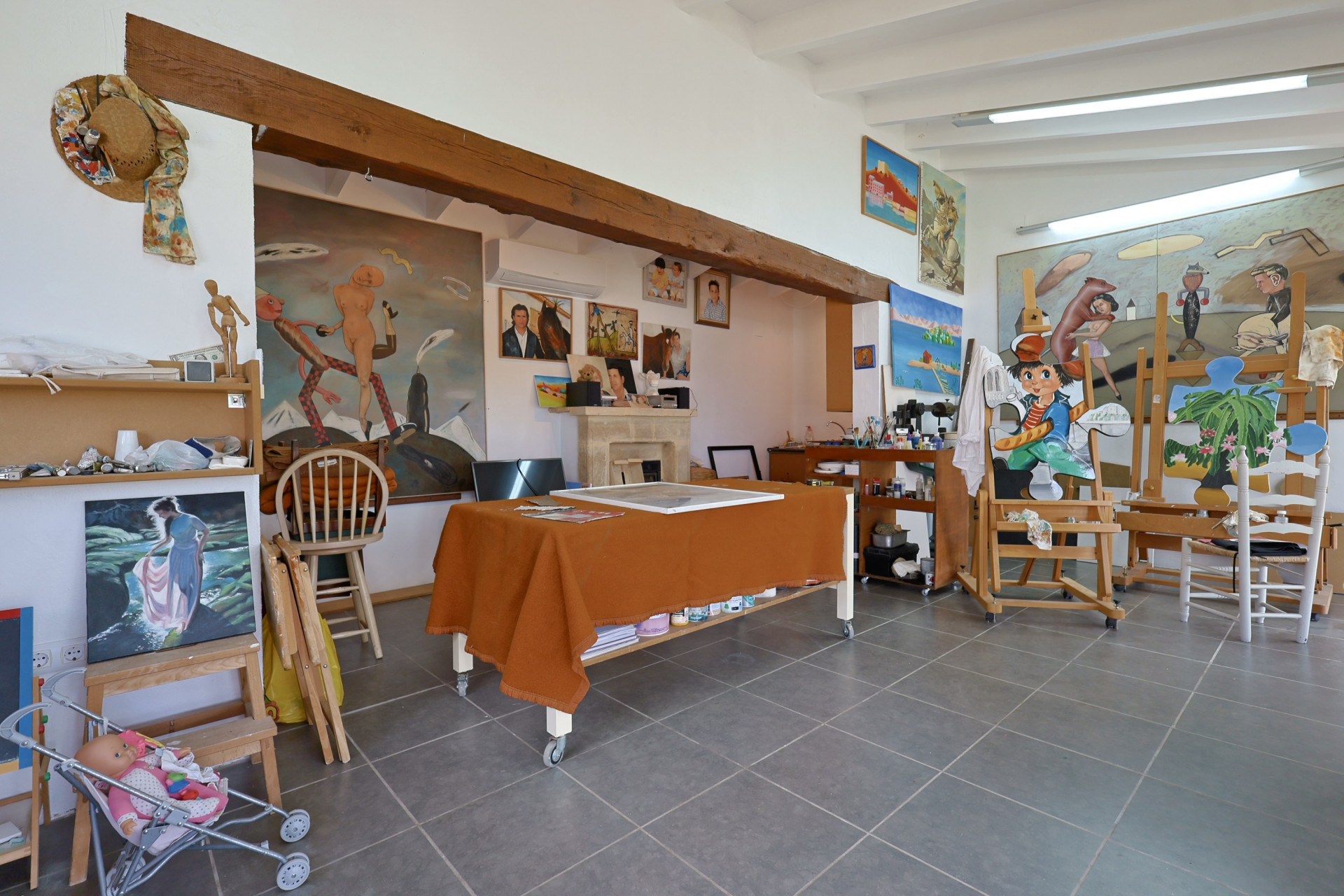 Atelier d'art spacieux avec sol carrelé gris, table recouverte d'un tissu orange et chevalets présentant des peintures.