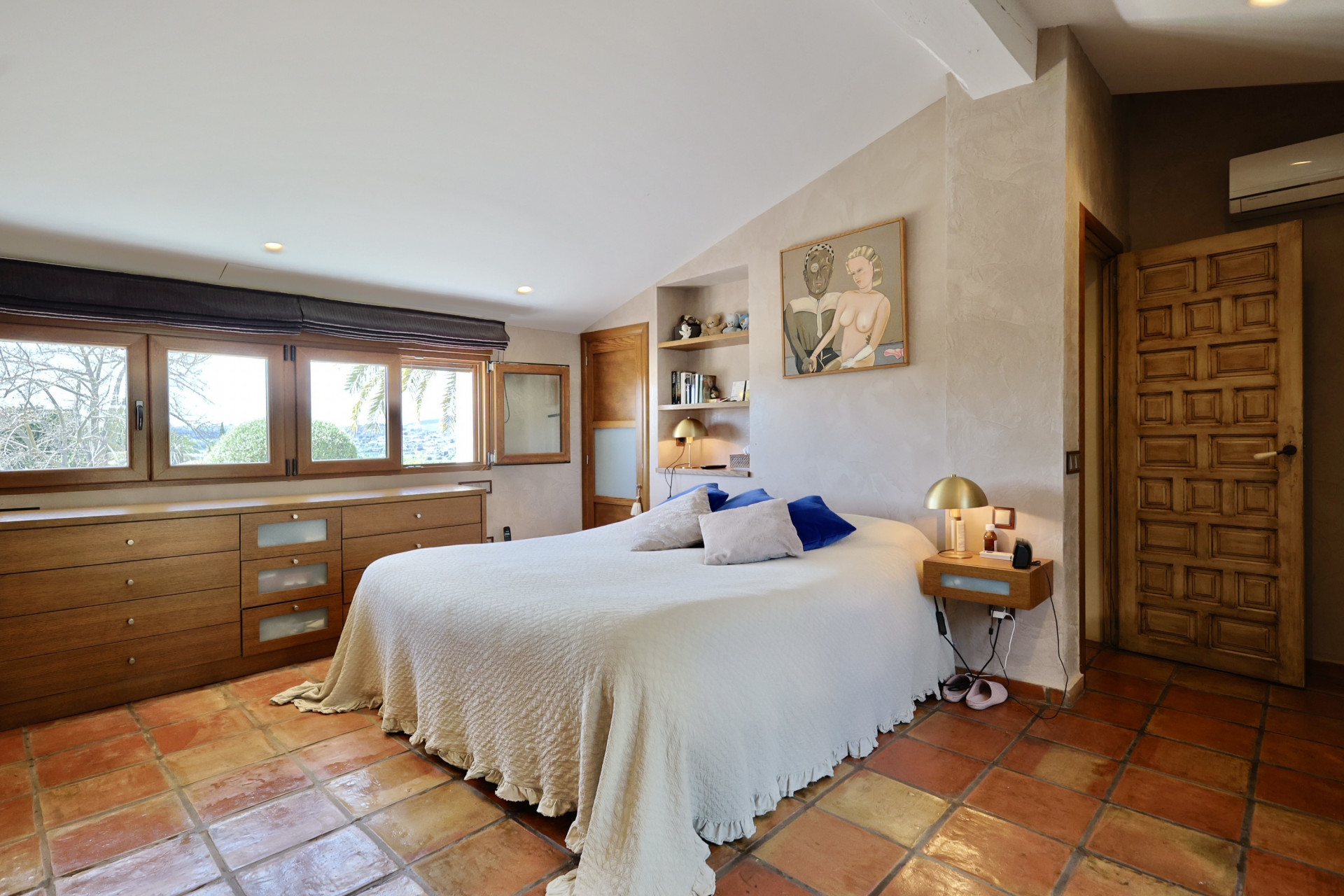 Chambre avec sol en terre cuite, lit double avec linge blanc et tête de lit en bois avec fenêtres.