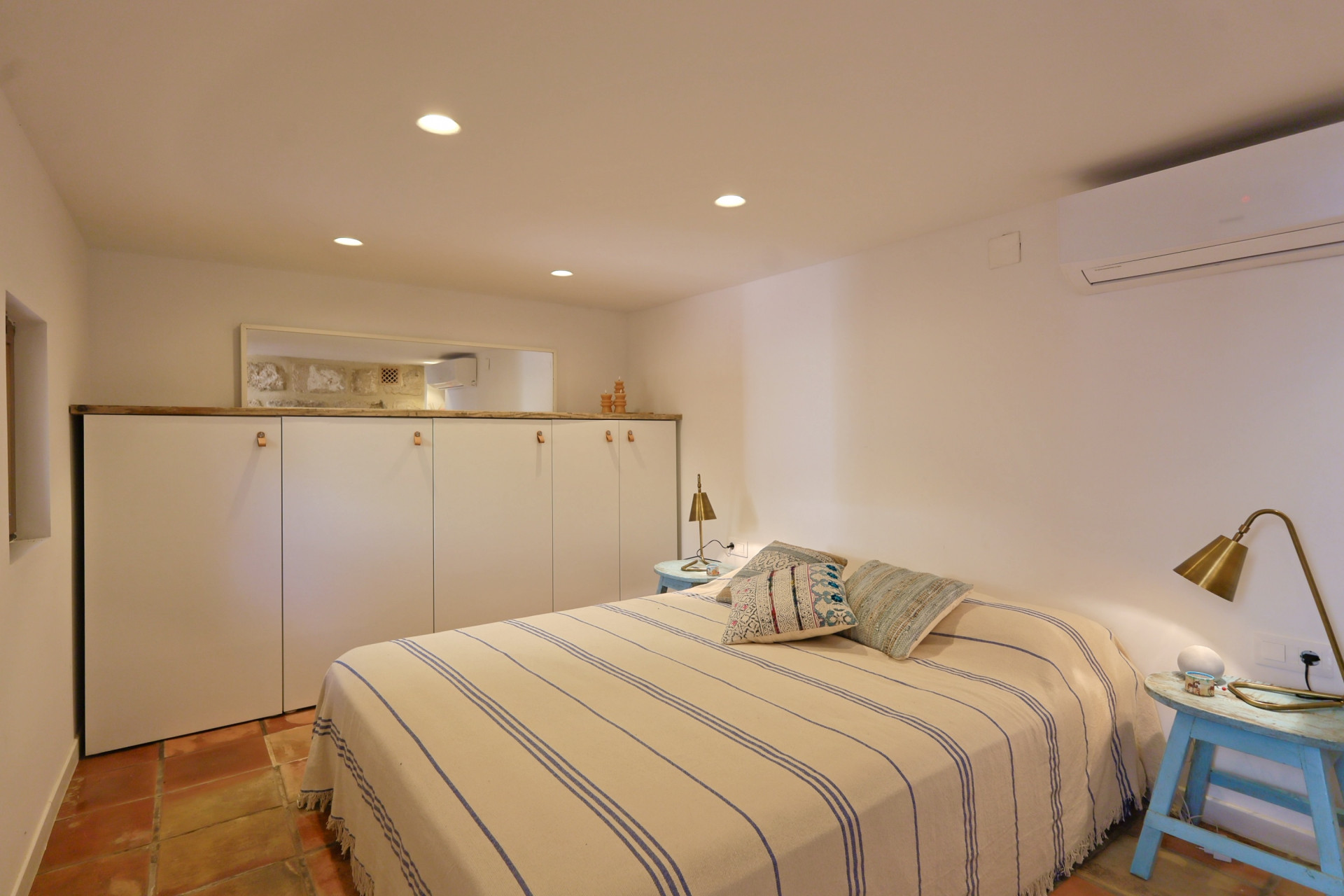 Chambre lumineuse avec sol en terre cuite, couvre-lit rayé et armoire intégrée blanche avec détails en bois.