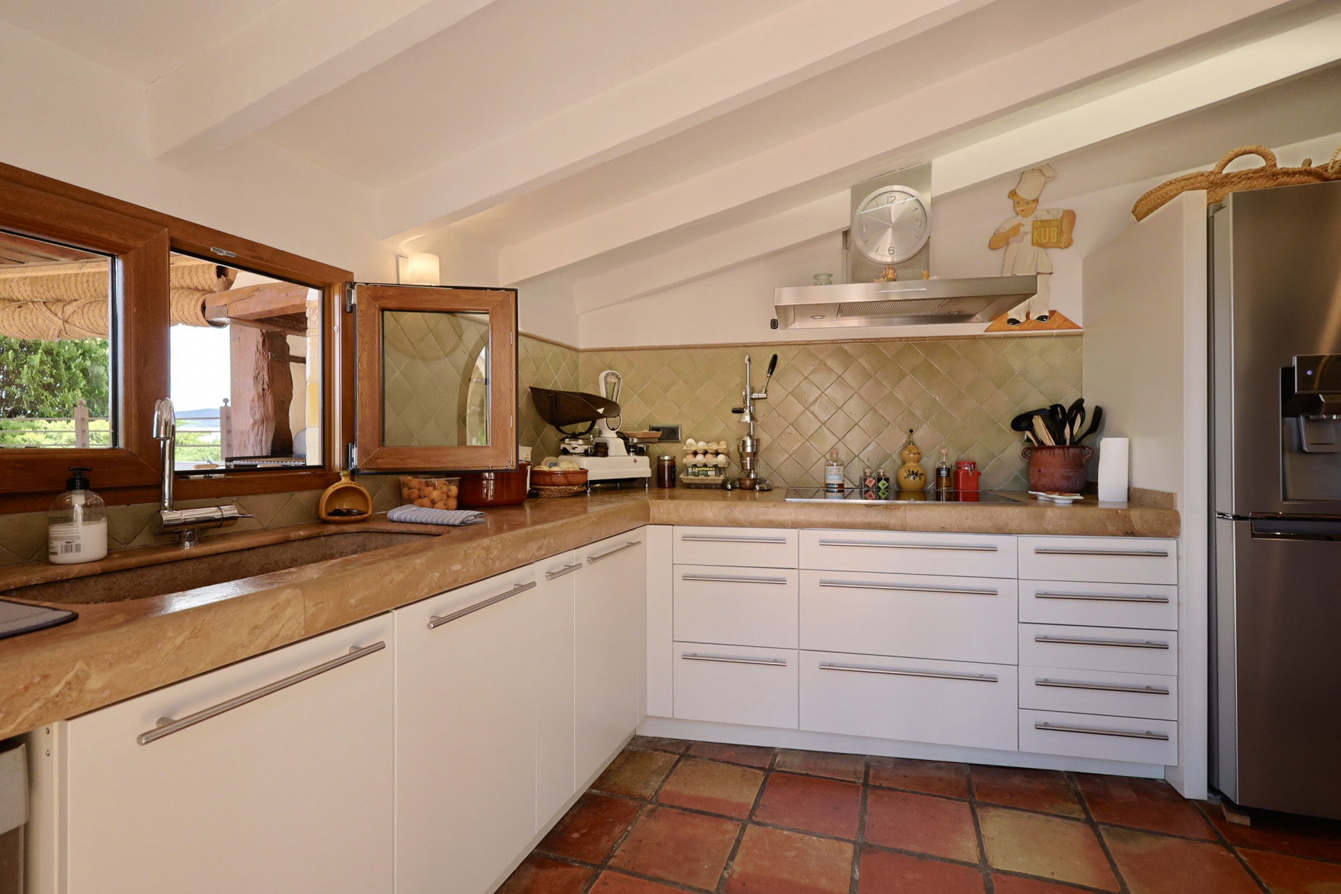 Cuisine rustique avec comptoir en pierre, armoires blanches et sol en terre cuite. Évier en pierre avec fenêtre en bois.