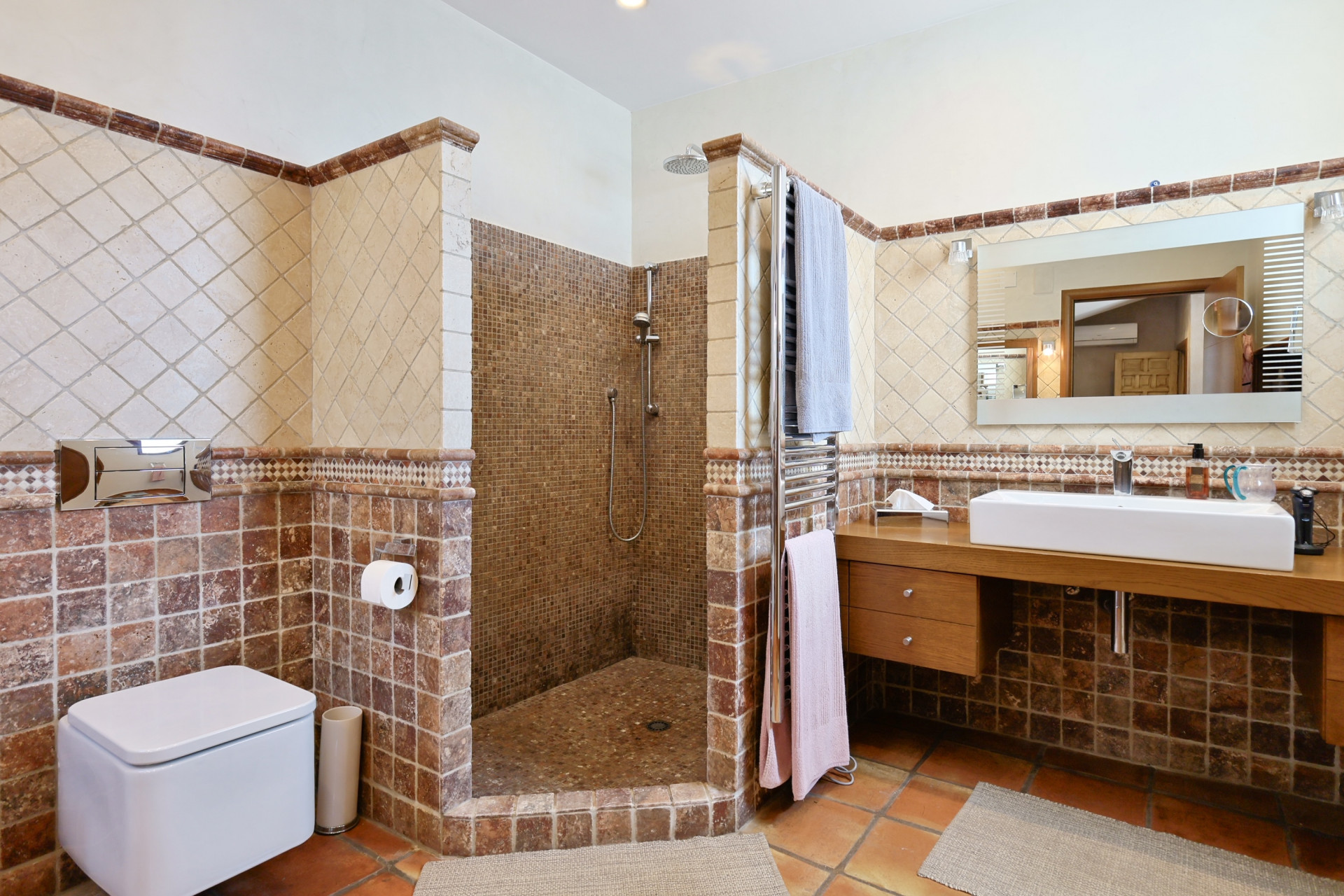 Détail de la salle de bain avec douche en mosaïque, lavabo moderne sur comptoir en bois et carrelage rustique.