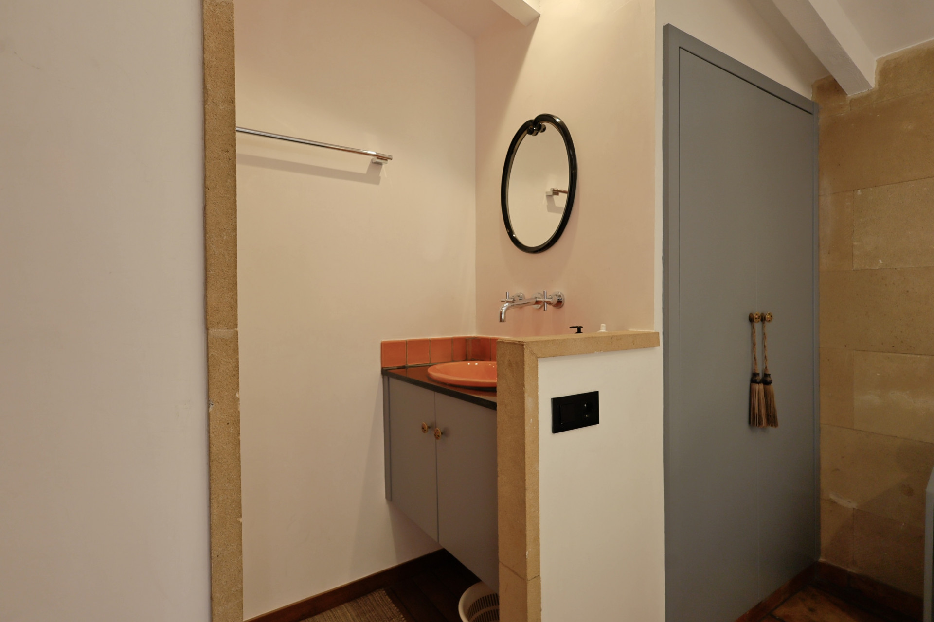 Détail de salle de bain avec lavabo en céramique orange, miroir ovale et porte grise à glands décoratifs.
