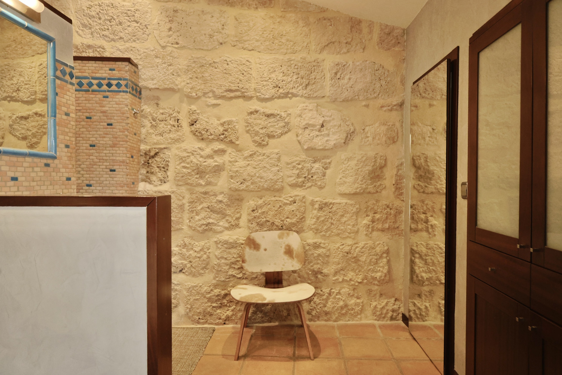 Détail de salle de bain avec mur en pierre rustique, miroir à cadre bleu et chaise design en peau de vache.