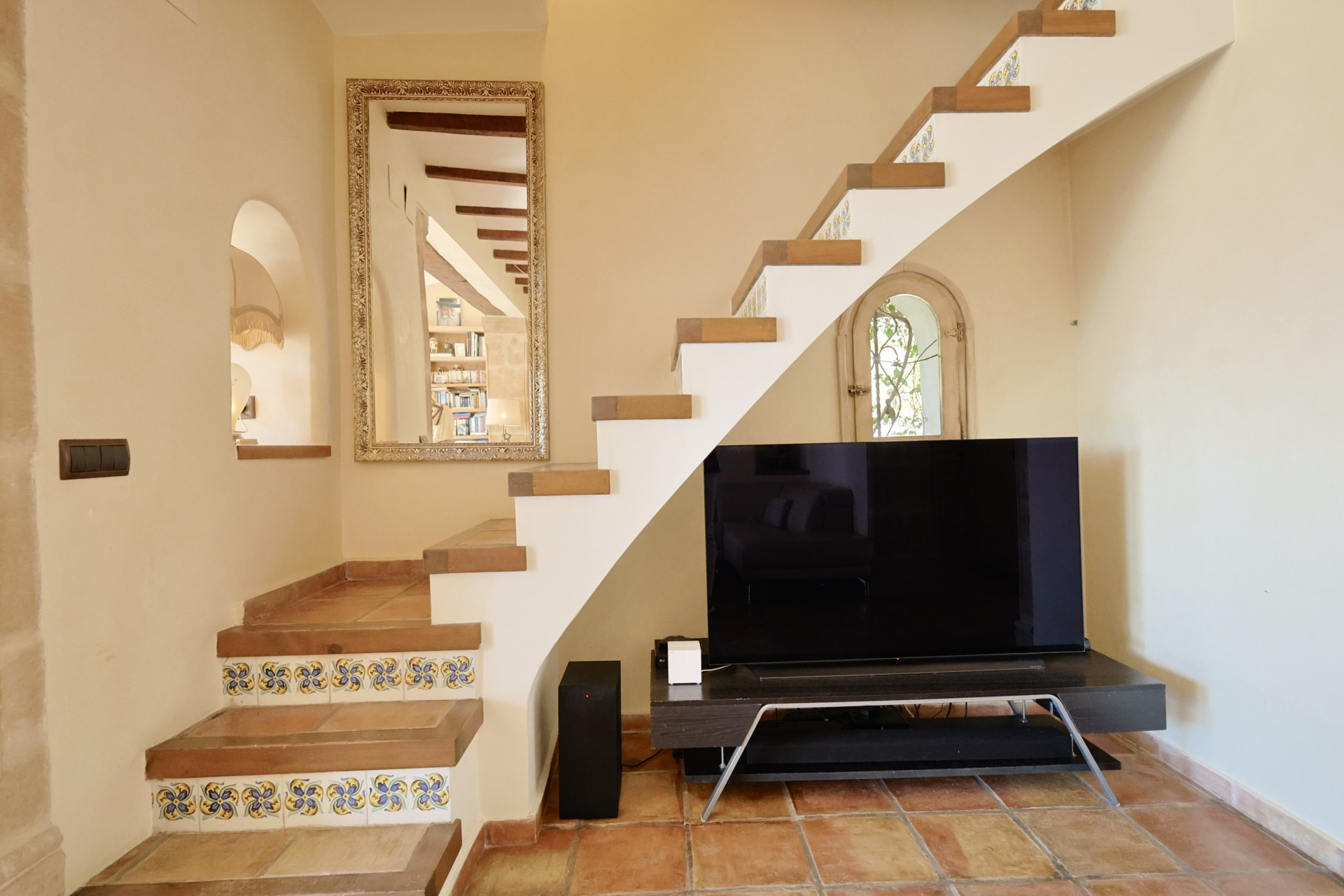 Détail d'escalier avec carreaux décoratifs et TV dans salon avec sol en terre cuite.