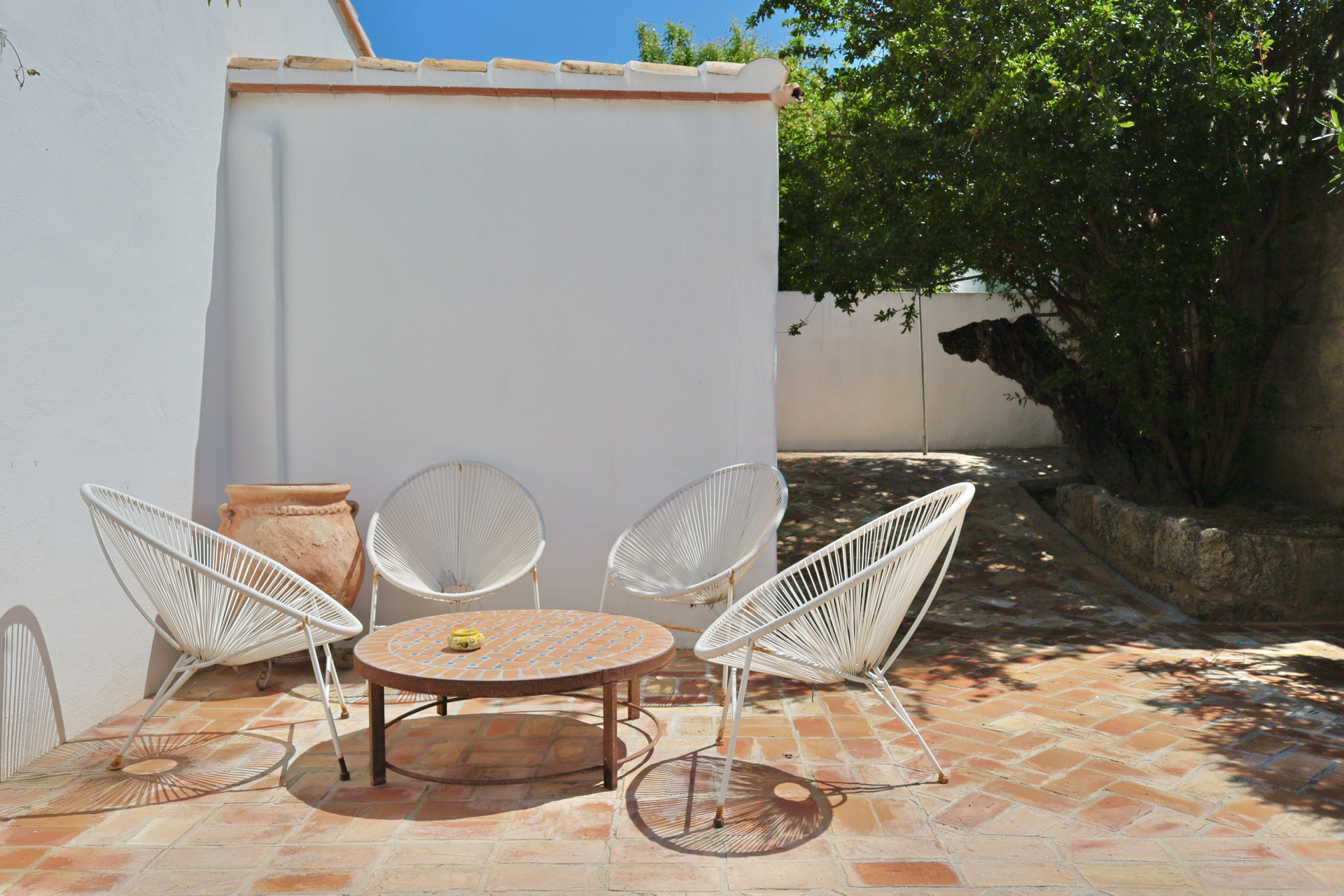 Patio avec sol en terre cuite, chaises en corde blanche et table en mosaïque, idéal pour la détente en plein air.