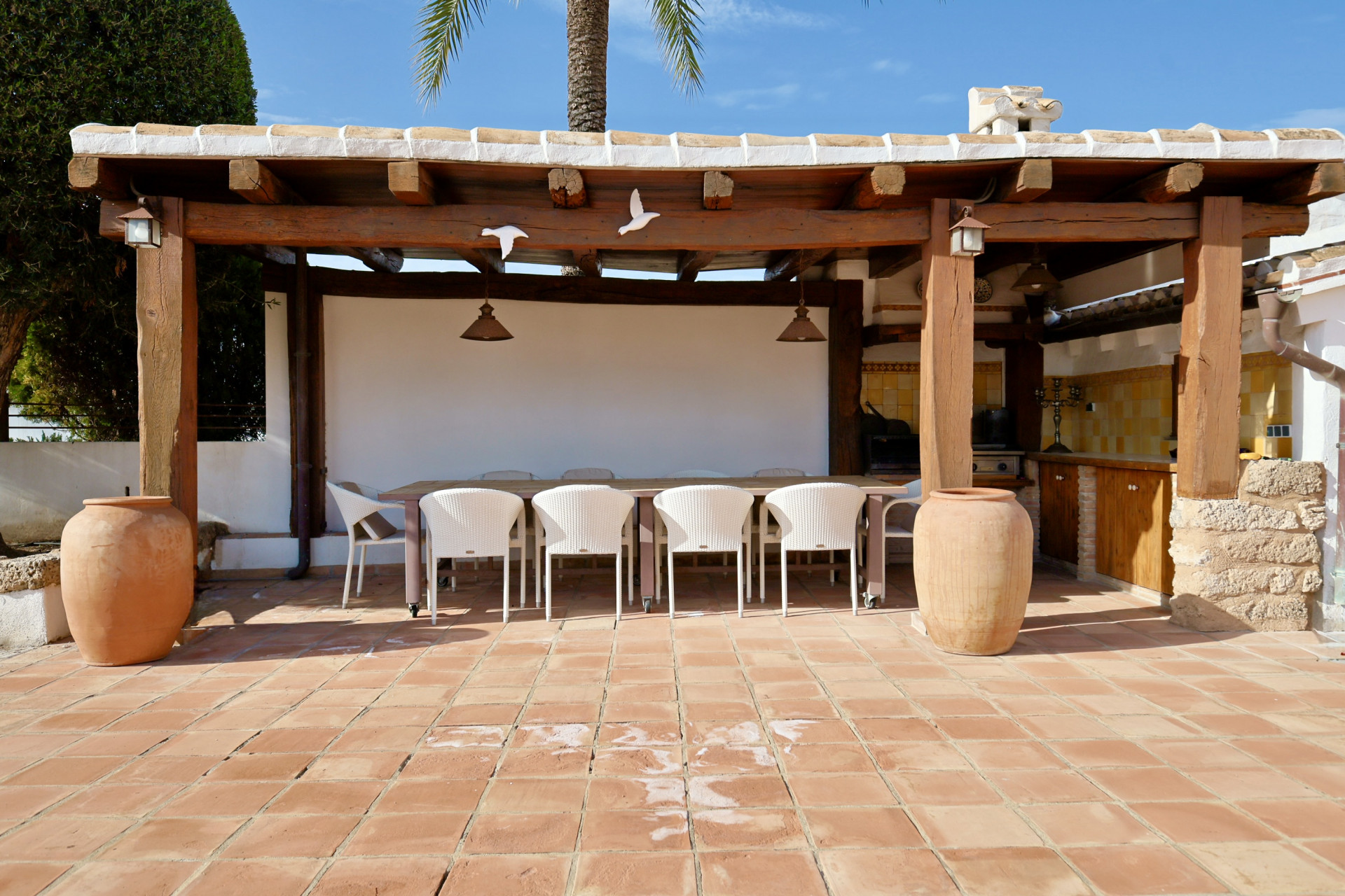 Salle à manger extérieure couverte avec longue table, chaises en osier blanches et sol en terre cuite.
