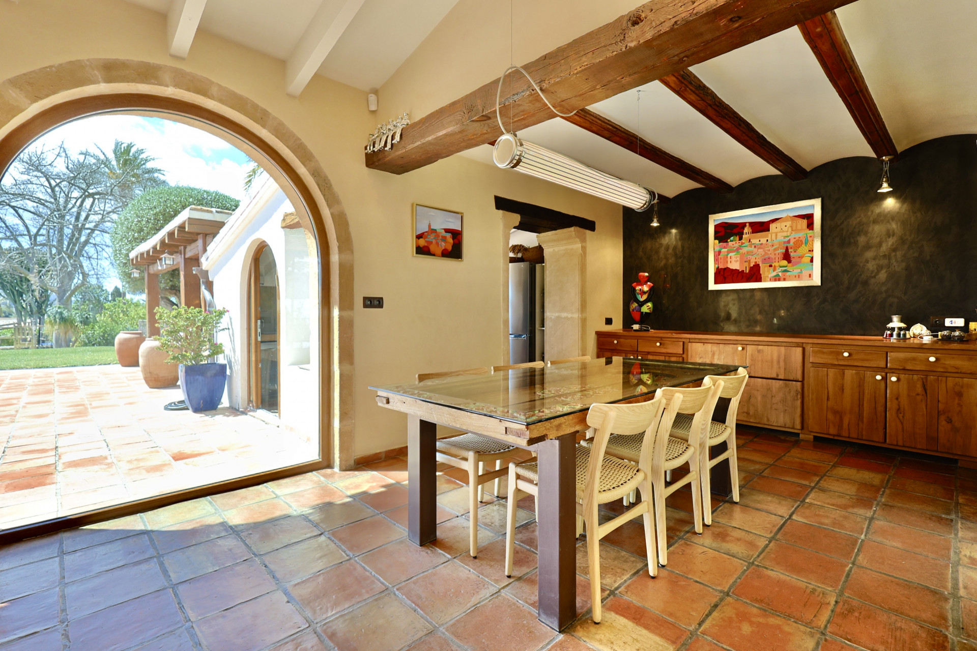Salle à manger rustique avec sol en terre cuite, table en bois et arche en pierre donnant sur le jardin.