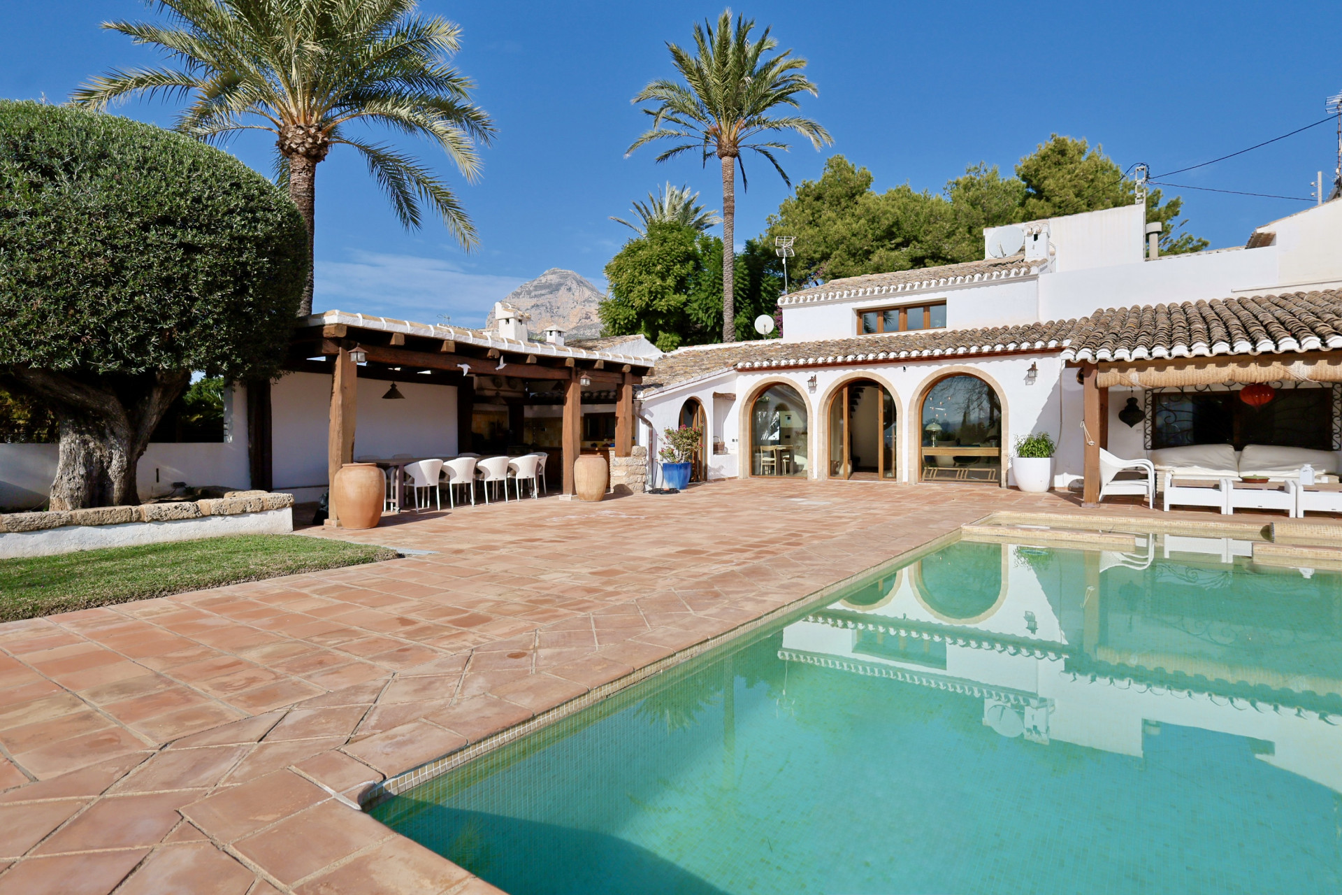 Vue de la piscine avec carrelage en terre cuite, terrasse couverte avec sièges et arcades de la villa.