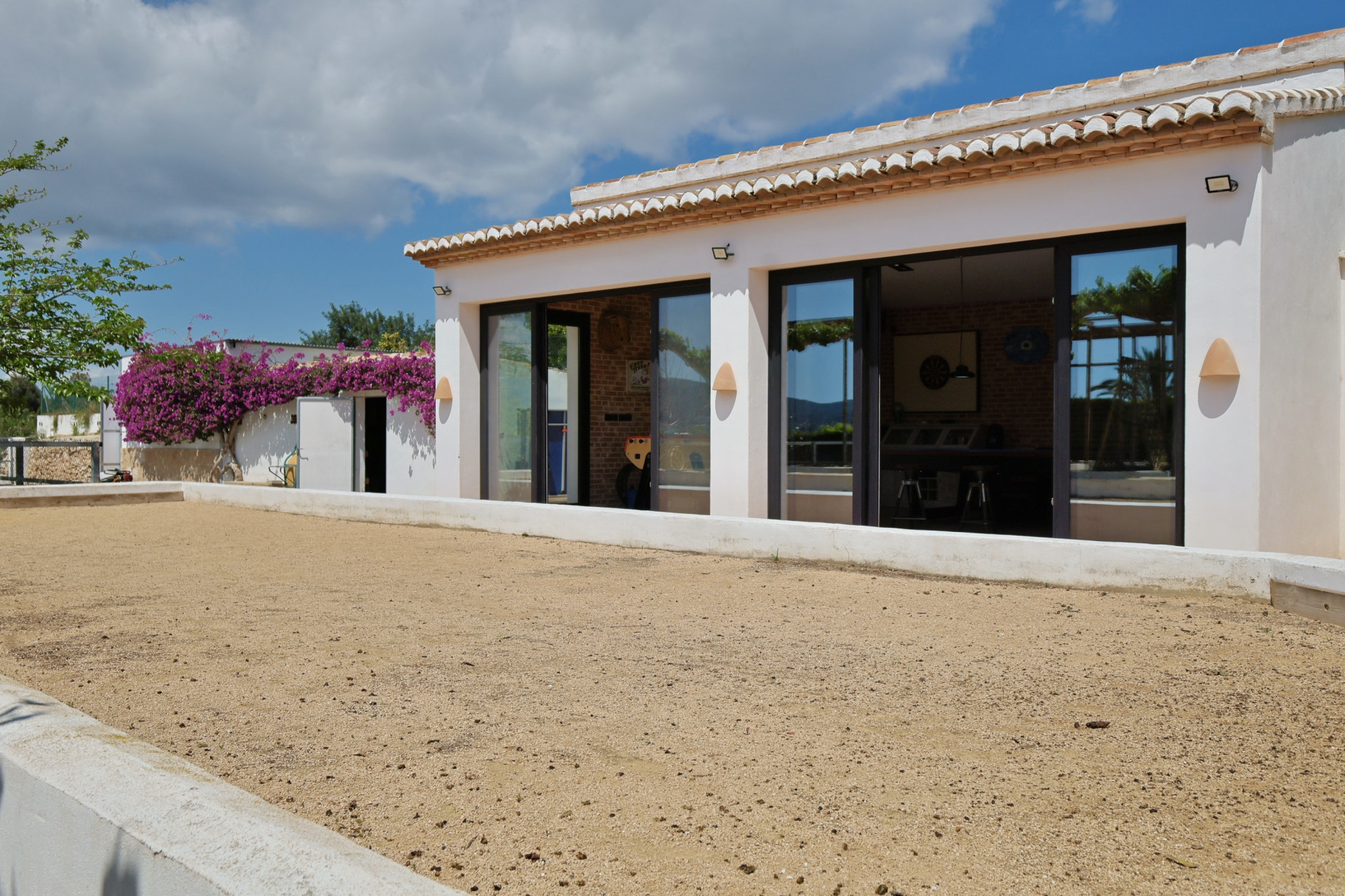 Vue extérieure de la villa avec terrain sablé et grandes fenêtres reflétant l'intérieur.