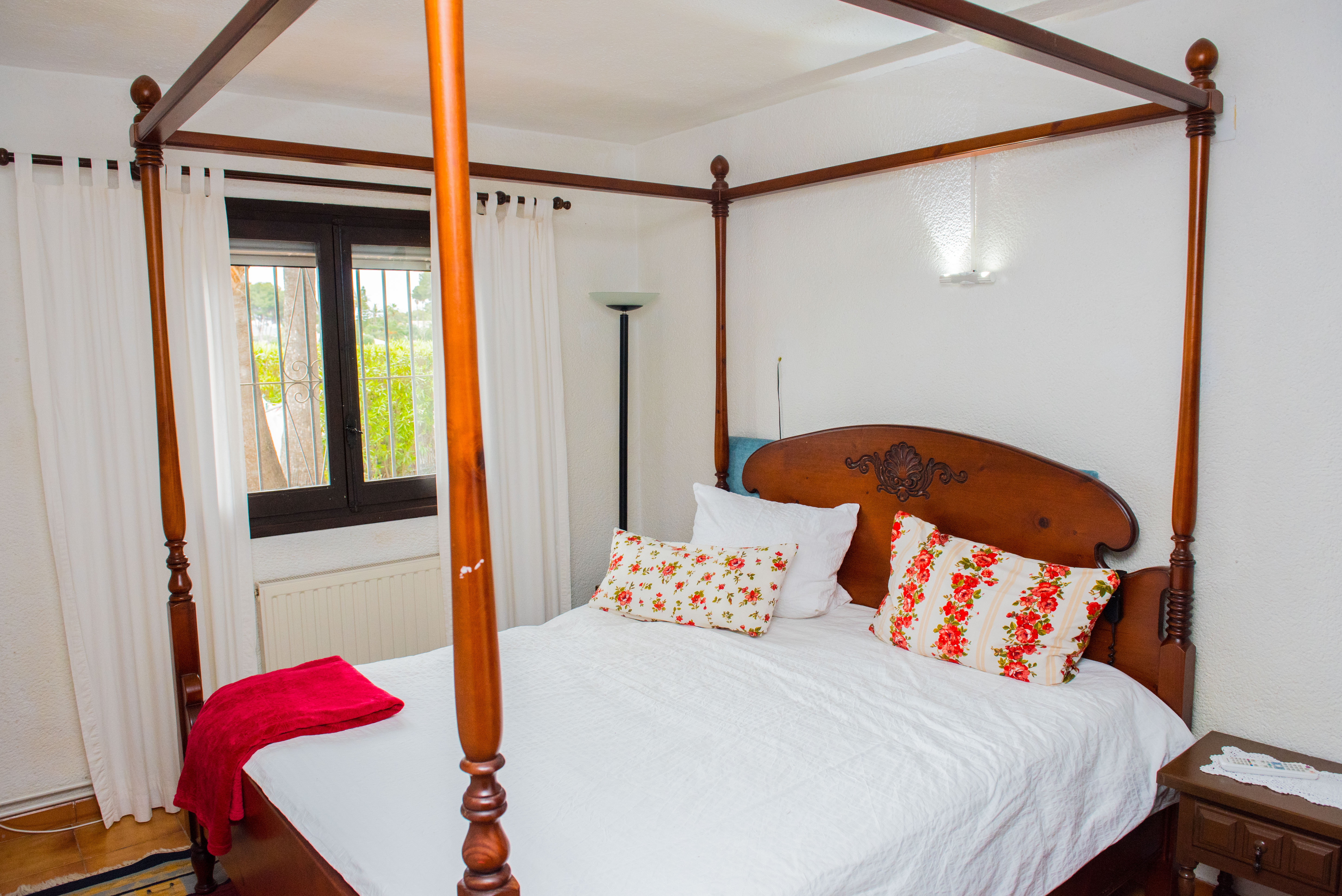 Chambre à coucher avec lit à baldaquin en bois sculpté, textiles à motifs floraux et fenêtre donnant sur le jardin avec radiateur.