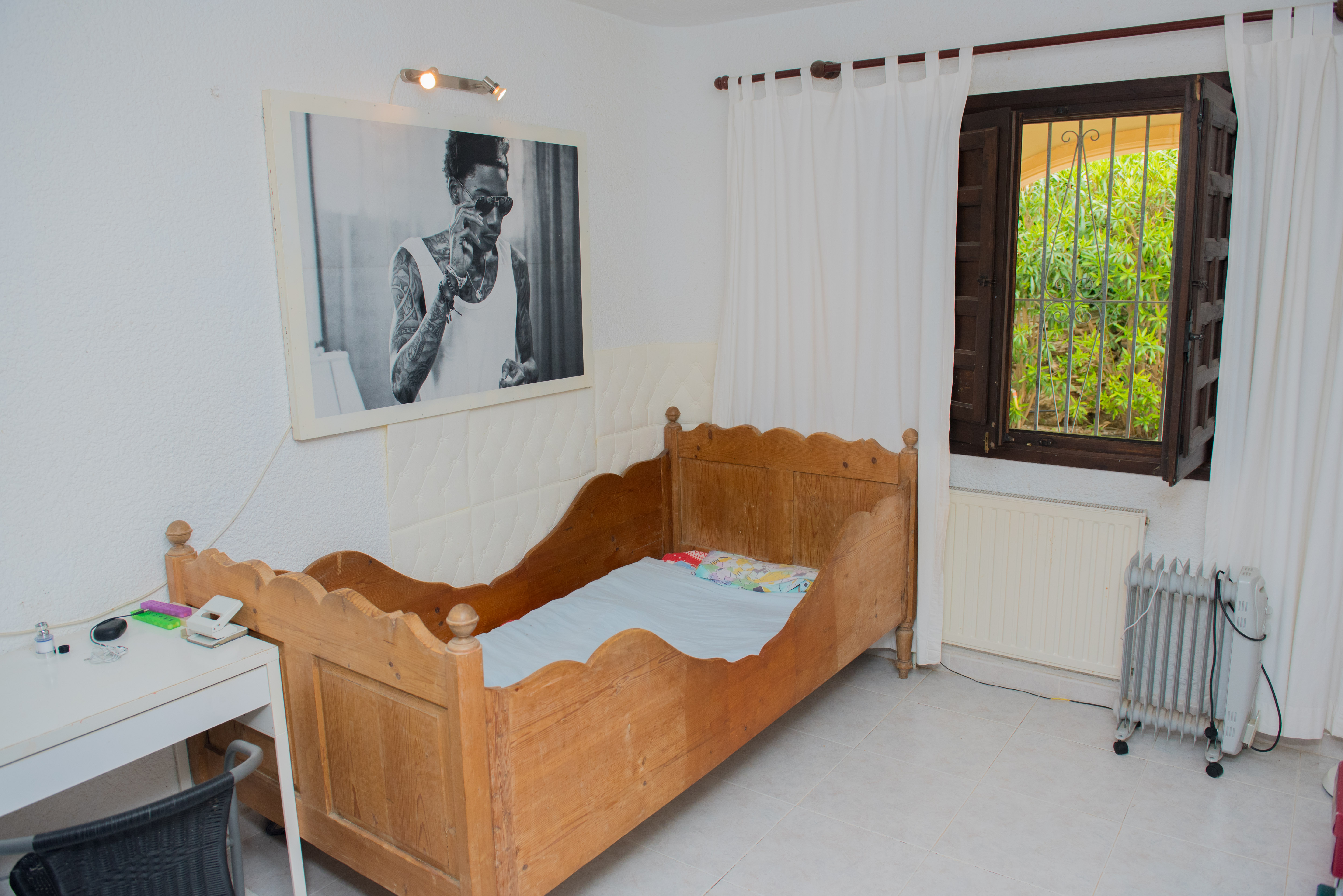 Chambre avec lit en bois rustique aux bords ondulés, bureau blanc et fenêtre en bois sombre. Sol en carrelage clair et vue sur la verdure environnante.