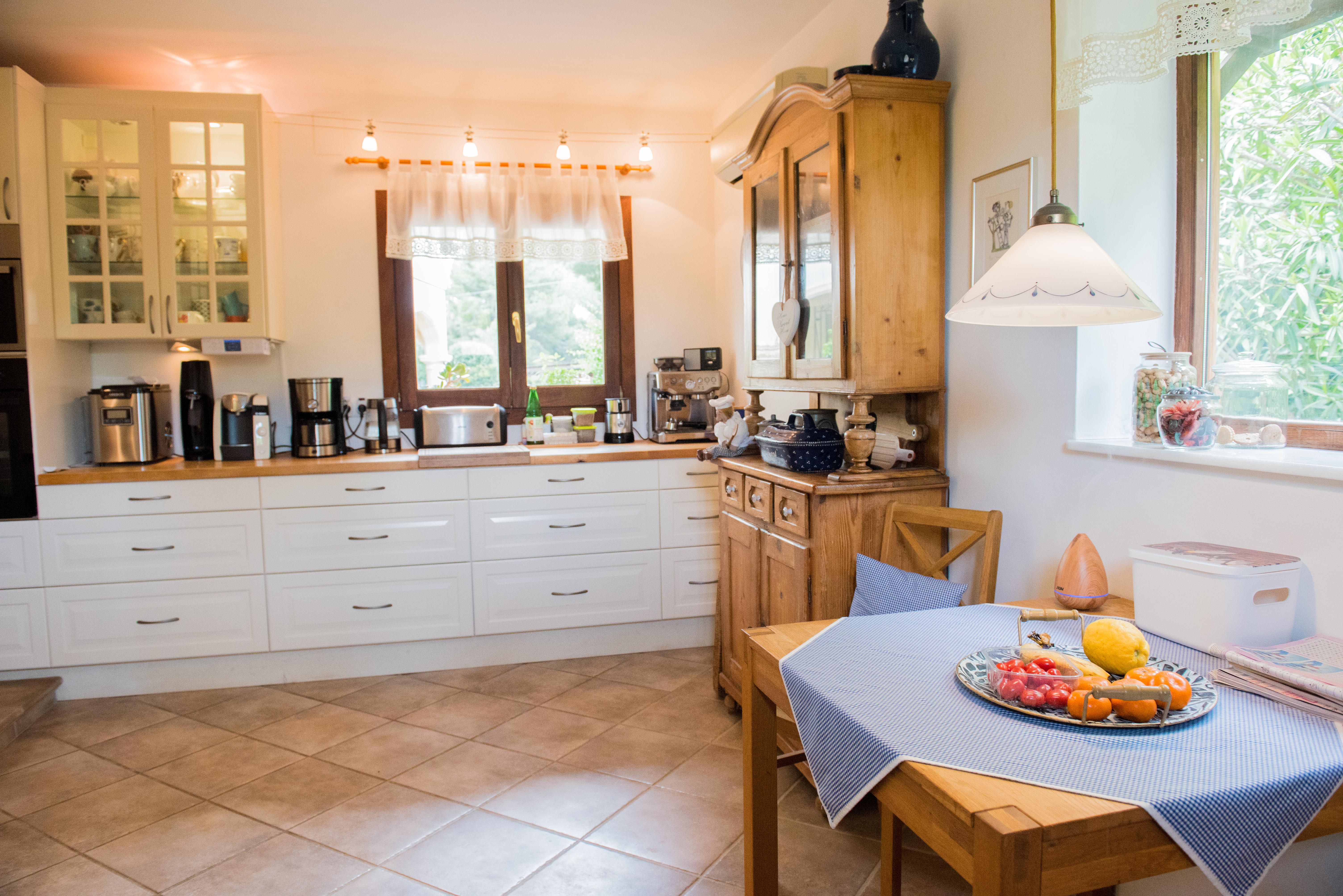 Cuisine rustique avec armoires blanches, plans de travail en bois et coin repas. Comprend un buffet en bois classique, un sol en céramique et la lumière naturelle de deux fenêtres.