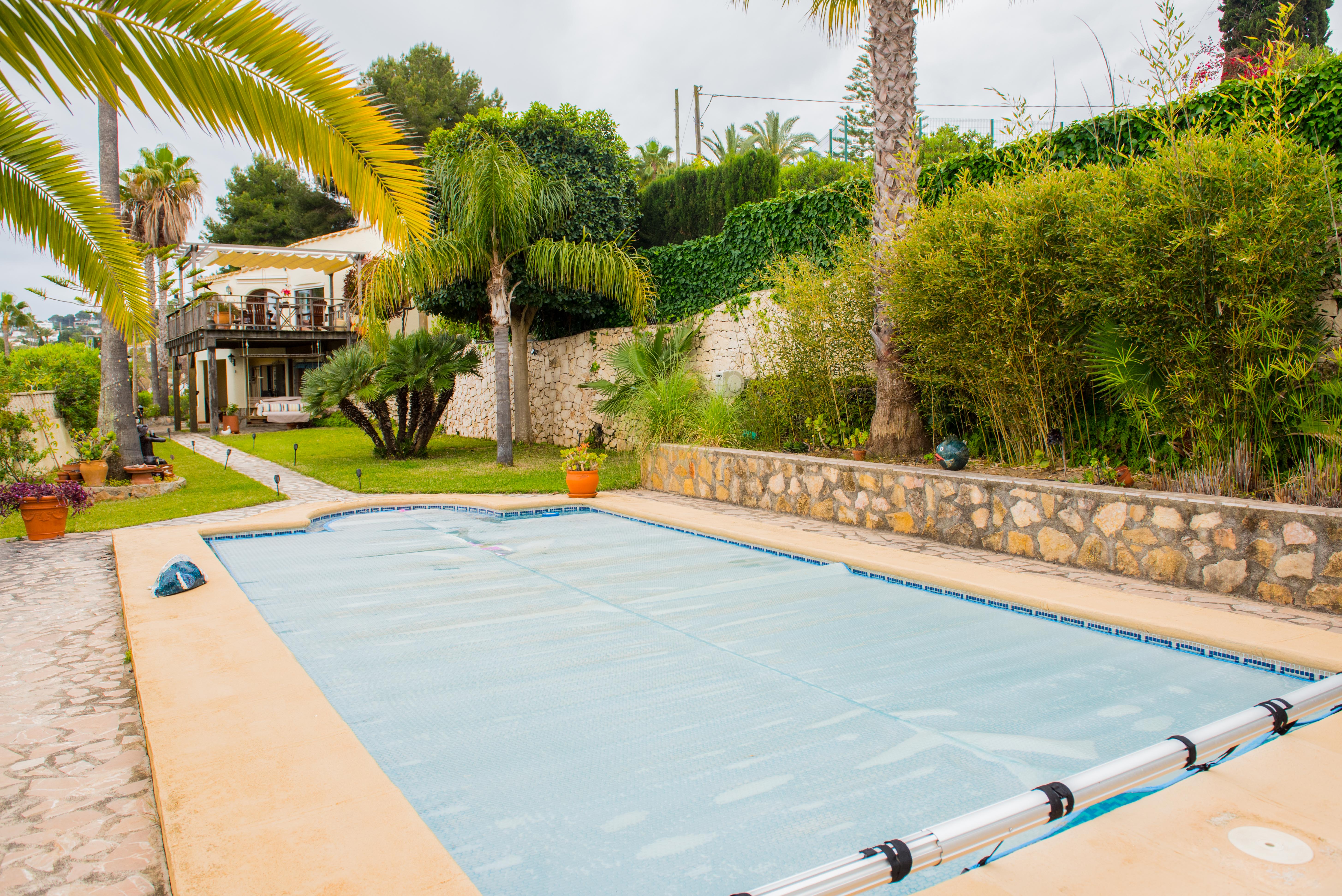 Piscine extérieure avec bâche thermique bleue et enrouleur, entourée de murs en pierre naturelle, de palmiers et d'un jardin méditerranéen luxuriant.