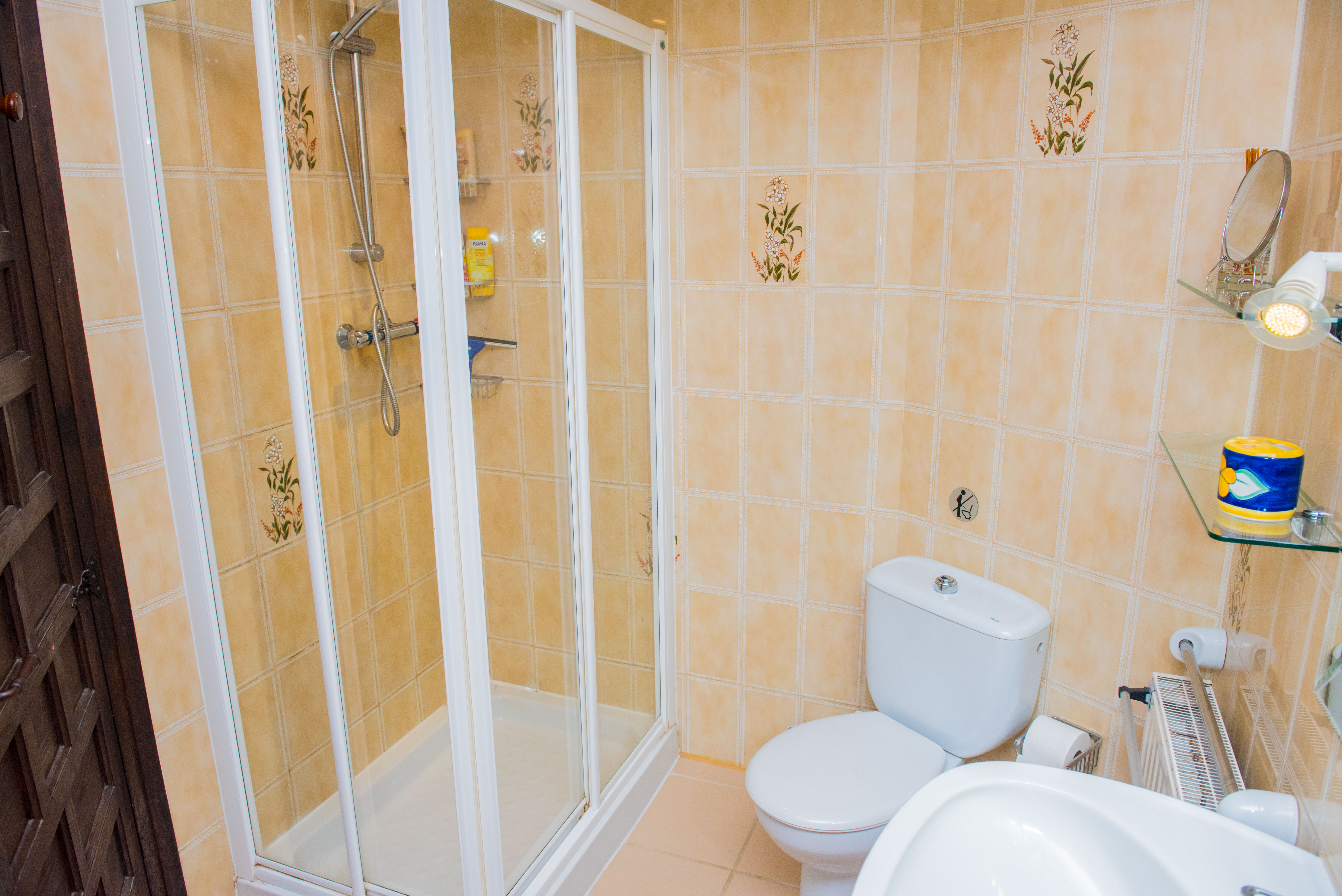 Salle de bain avec cabine de douche à cadre blanc, murs carrelés dans les tons beige avec motifs floraux et sanitaires en céramique blanche.