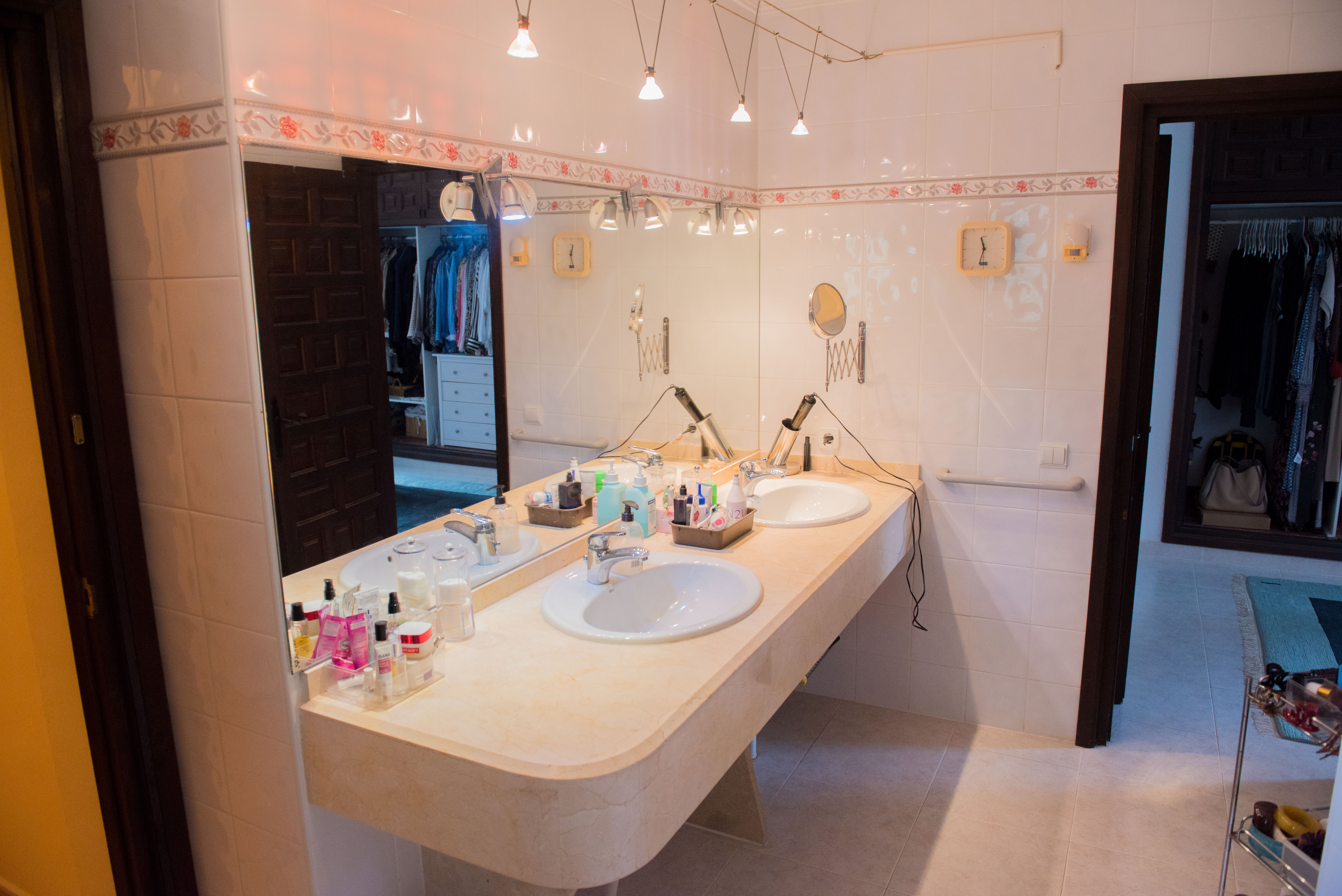 Salle de bain avec double vasque sur plan en marbre crème, grand miroir et carrelage blanc orné d'une frise florale, ouvrant sur un dressing.