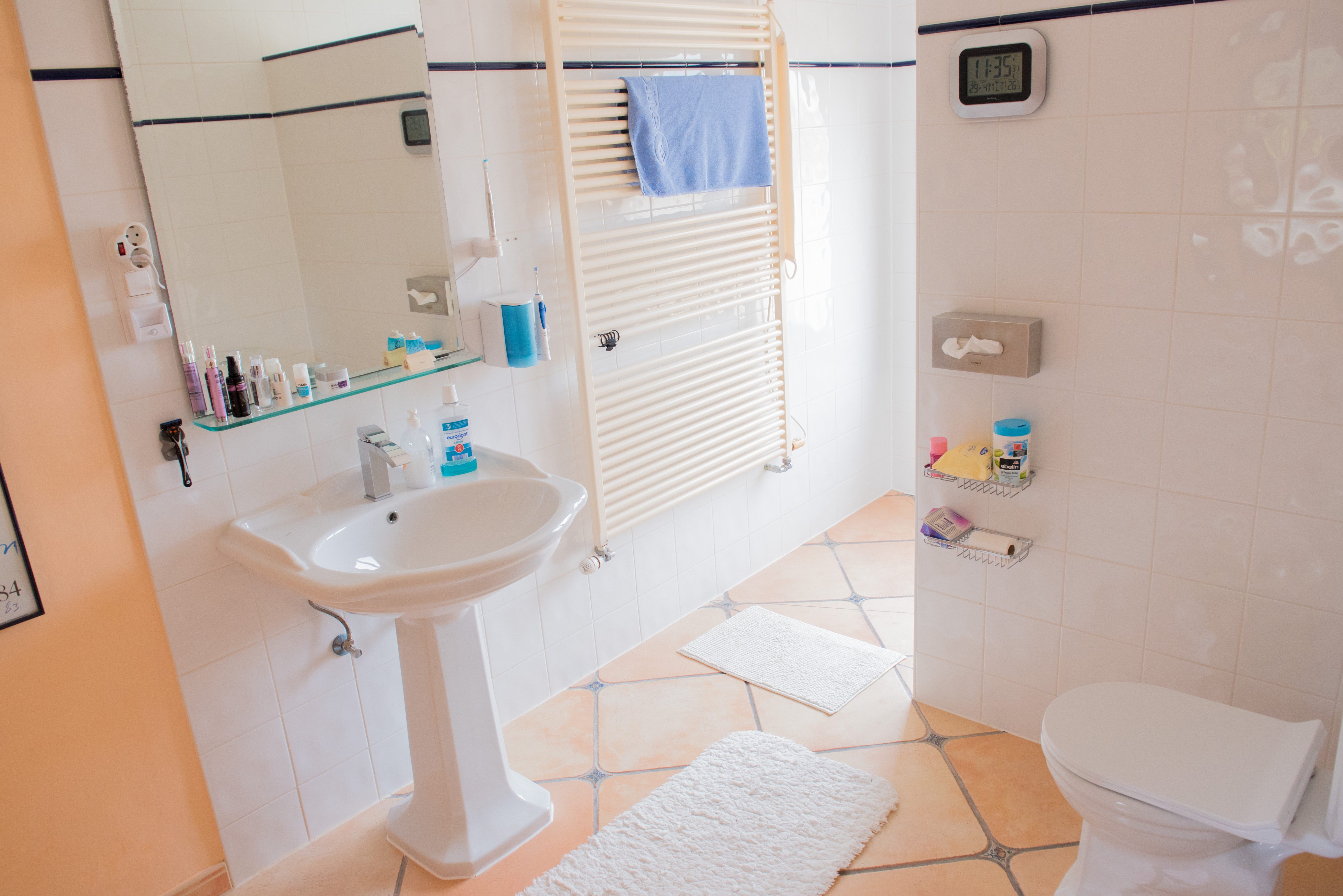 Salle de bain avec lavabo sur colonne, sol en terre cuite à cabochons, radiateur sèche-serviettes et murs carrelés blancs soulignés d'un liseré bleu.