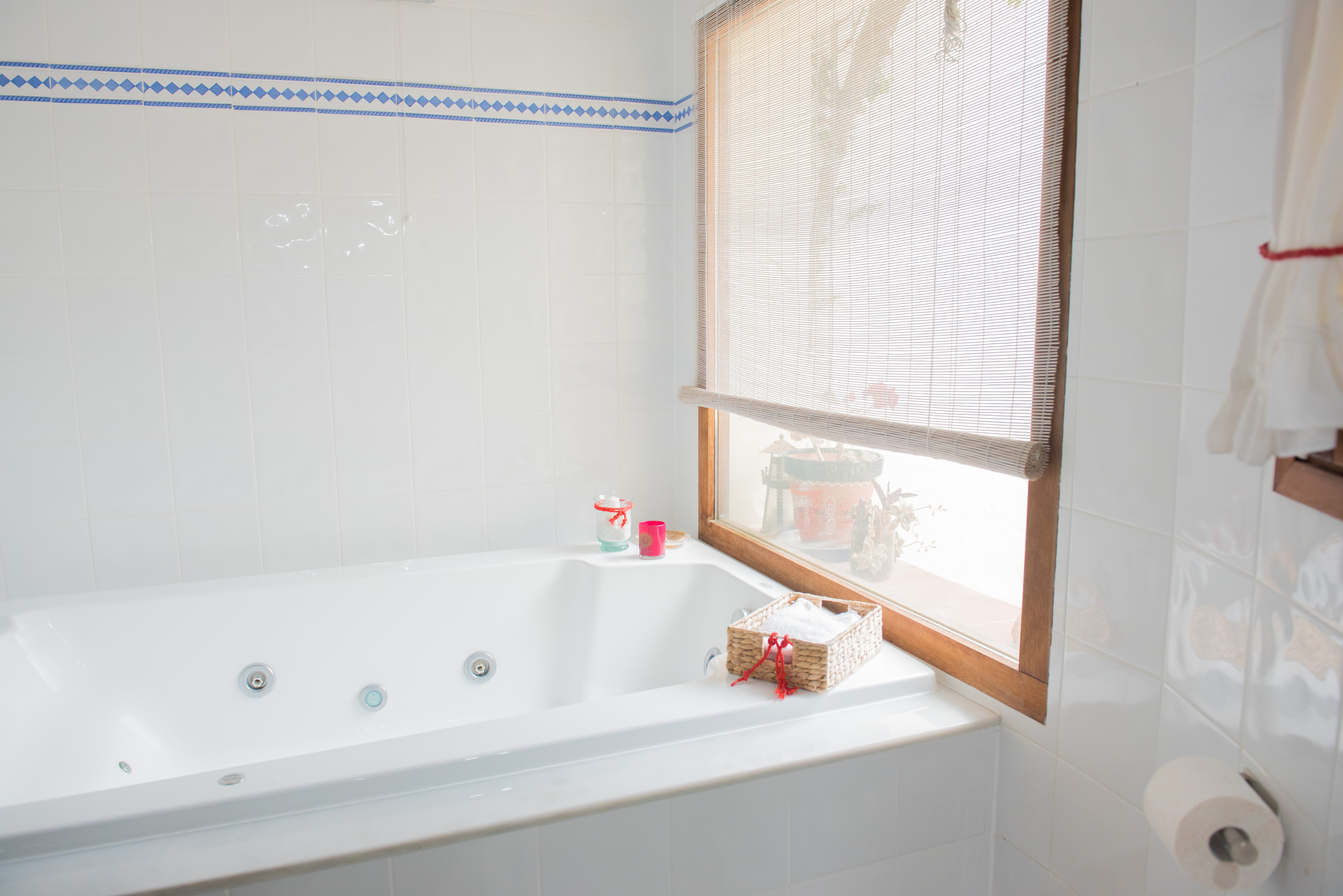 Salle de bain équipée d'une baignoire balnéo, de murs carrelés blancs avec frise bleue et d'une fenêtre en bois avec store enrouleur en bambou.