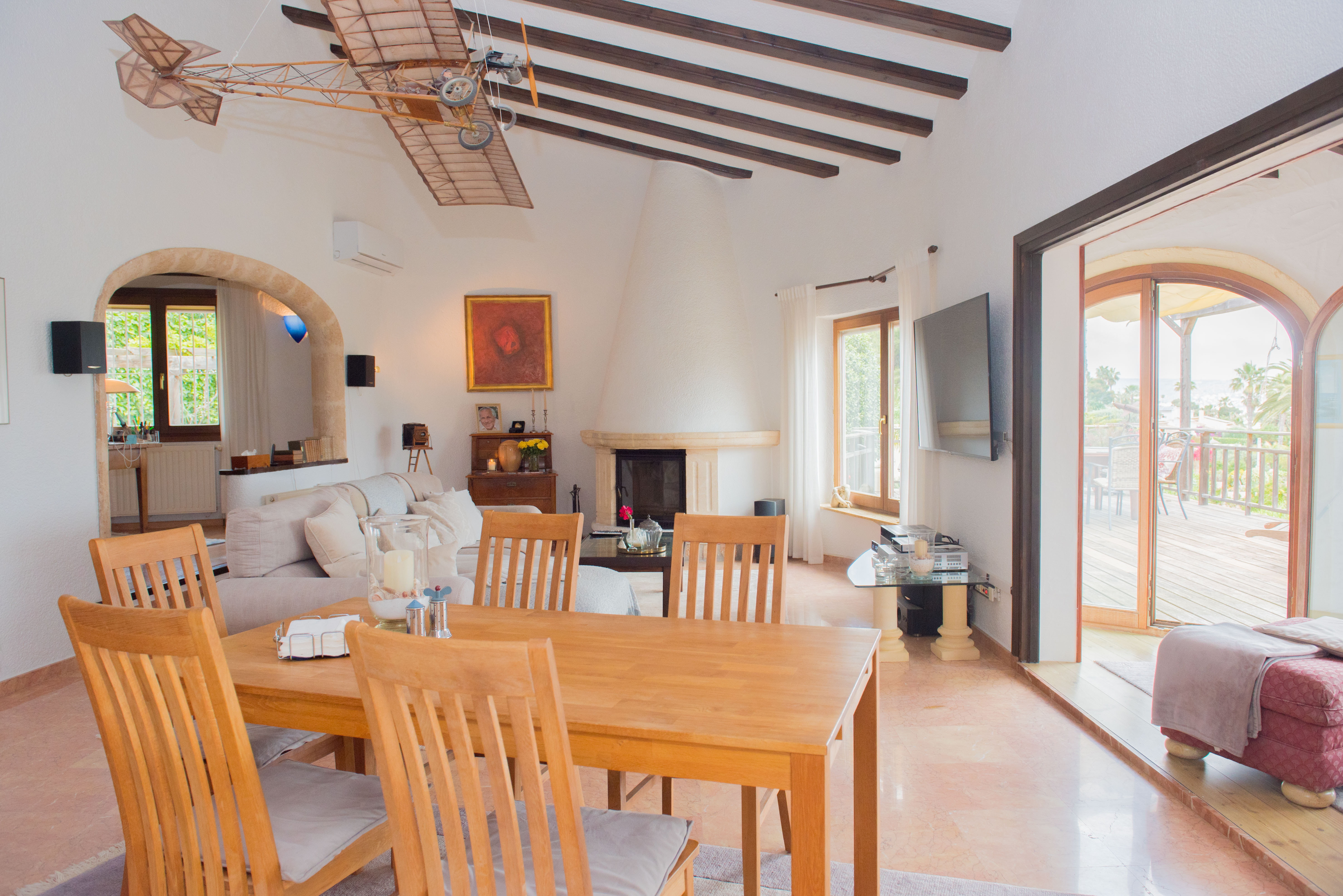 Salon-salle à manger avec poutres apparentes, cheminée conique blanche et accès direct à la terrasse en bois par de grandes baies vitrées cintrées.