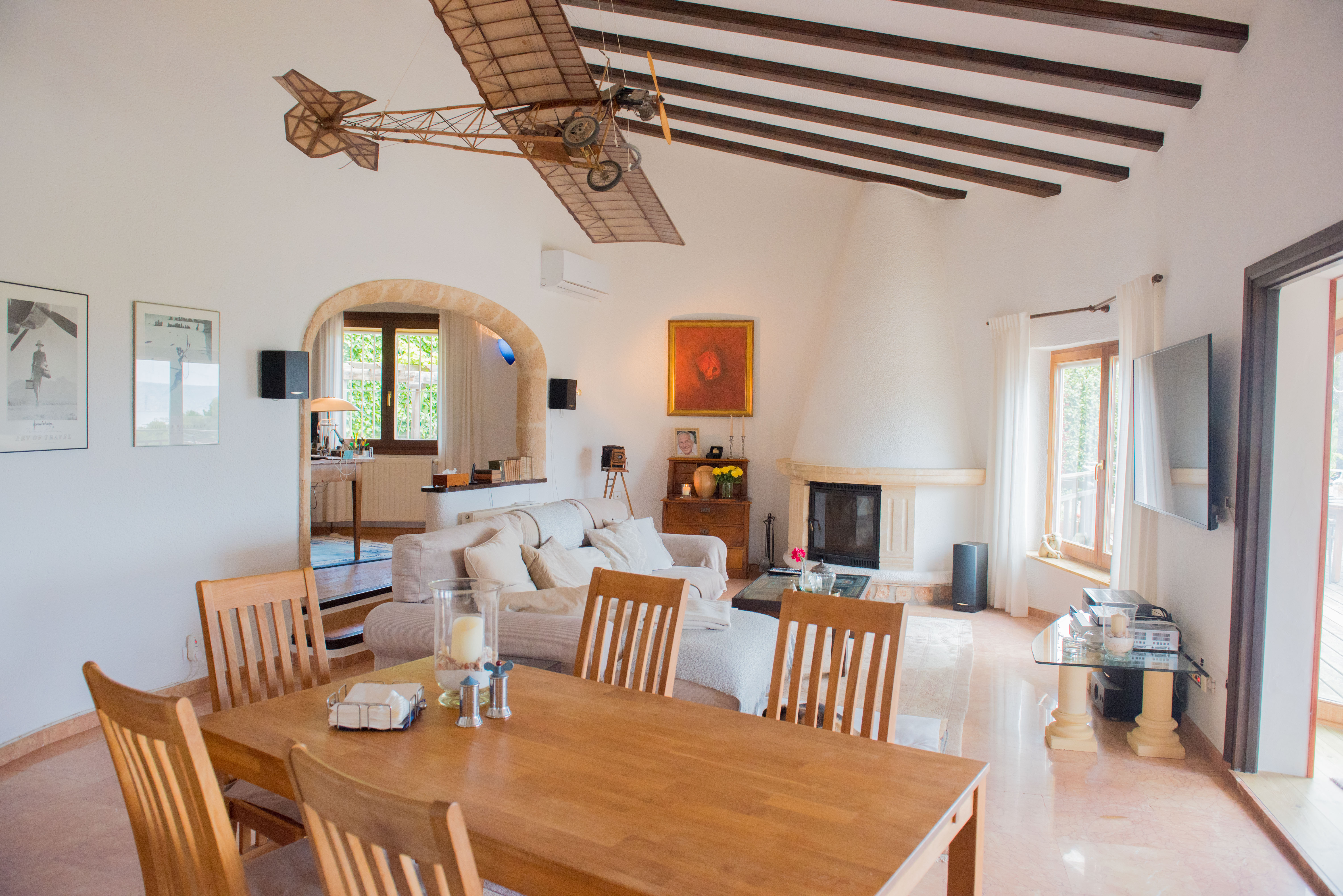 Salon-salle à manger avec poutres apparentes, cheminée conique blanche et arche en pierre. L'espace offre de hauts plafonds et un accès direct au bureau attenant.