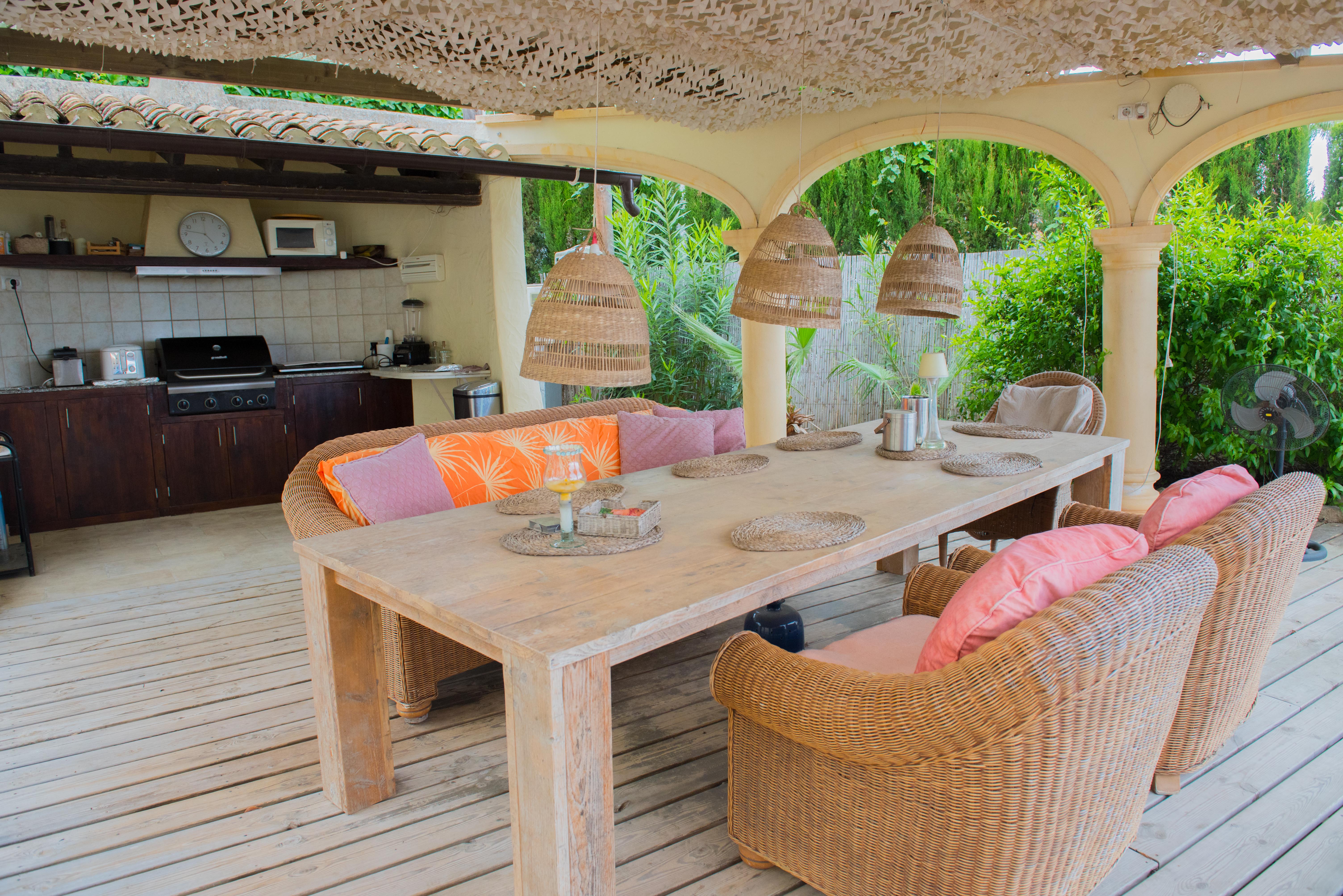 Terrasse couverte avec cuisine d'été équipée, table à manger en bois massif, fauteuils en osier et ombrage décoratif sur un sol en bois.