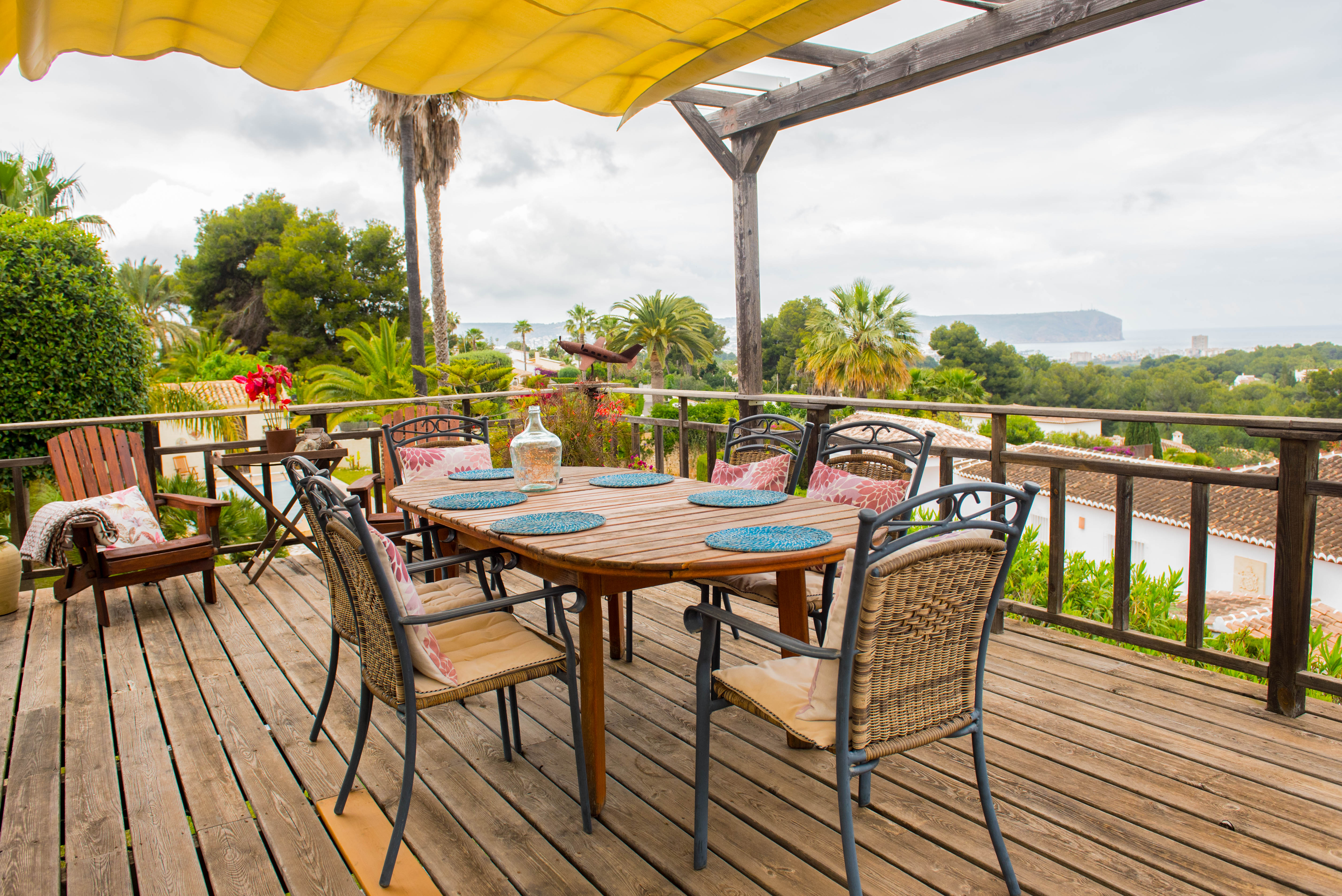 Terrasse en bois avec store banne jaune, comprenant un coin repas extérieur et une vue panoramique sur la mer et les environs.