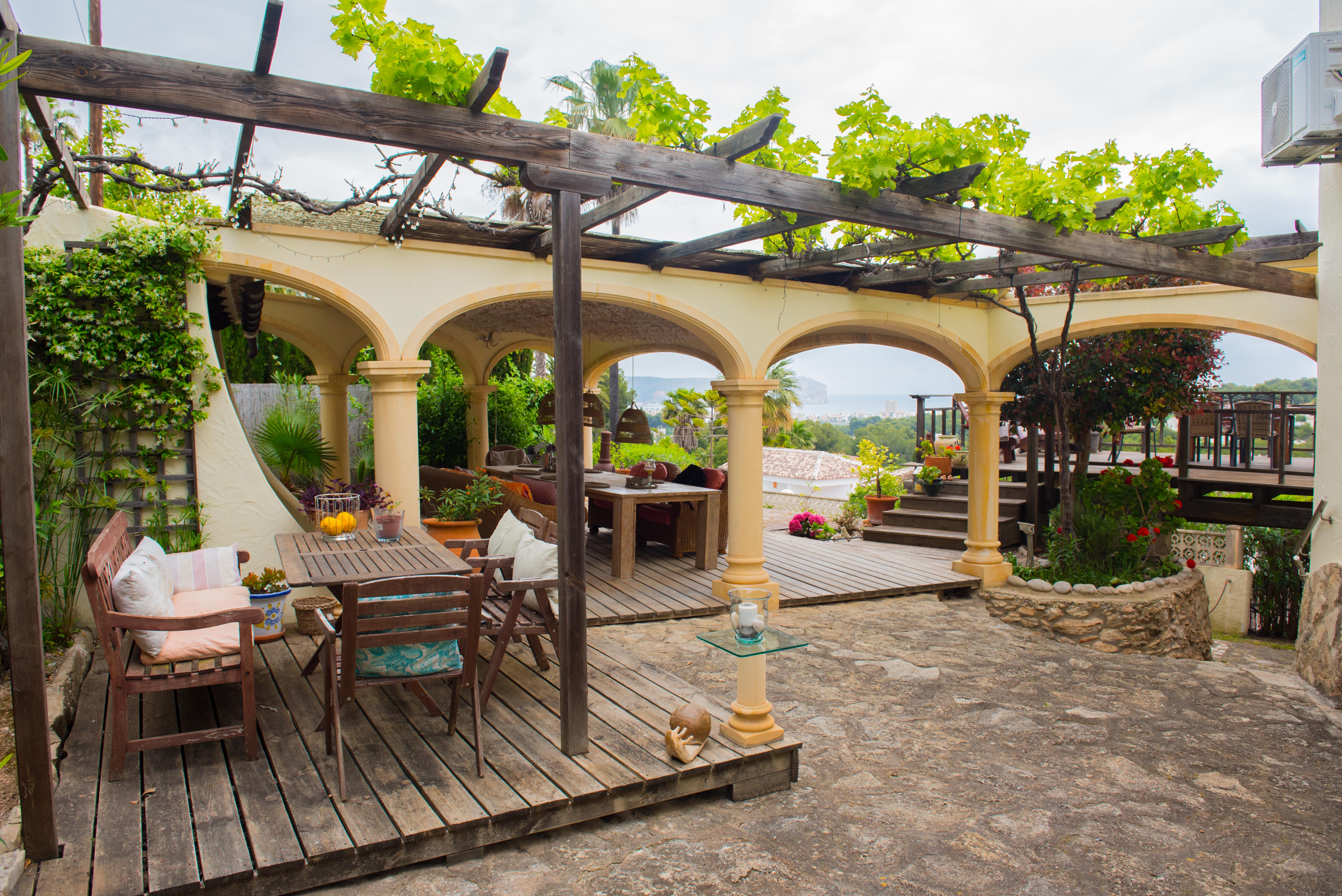 Terrasse extérieure avec pergola en bois et vignes, plateforme en bois et porche à arcades offrant une vue panoramique sur la mer et la côte.