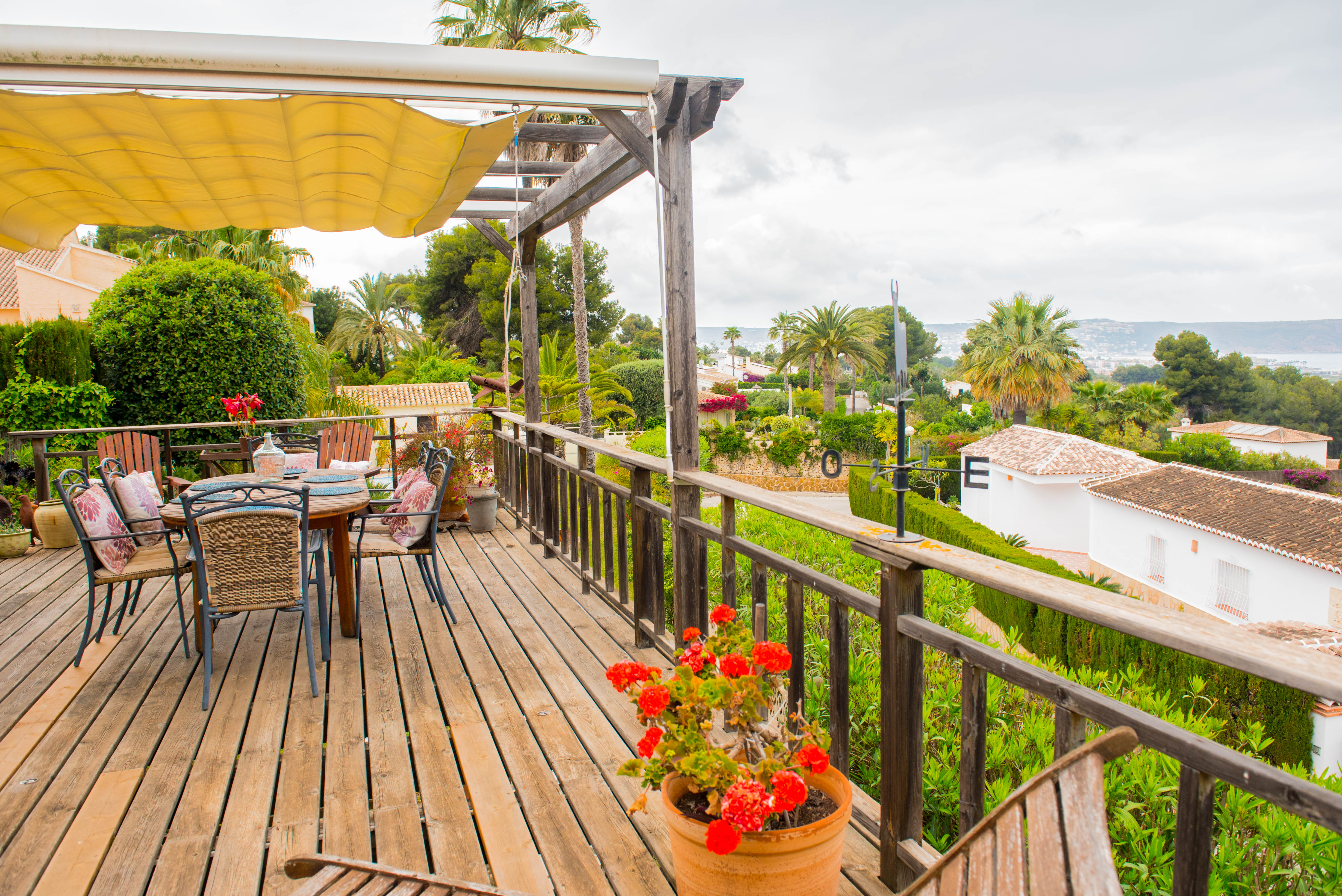 Terrasse surélevée en bois avec store banne, coin repas extérieur et garde-corps rustique offrant une vue panoramique sur la mer et la végétation.