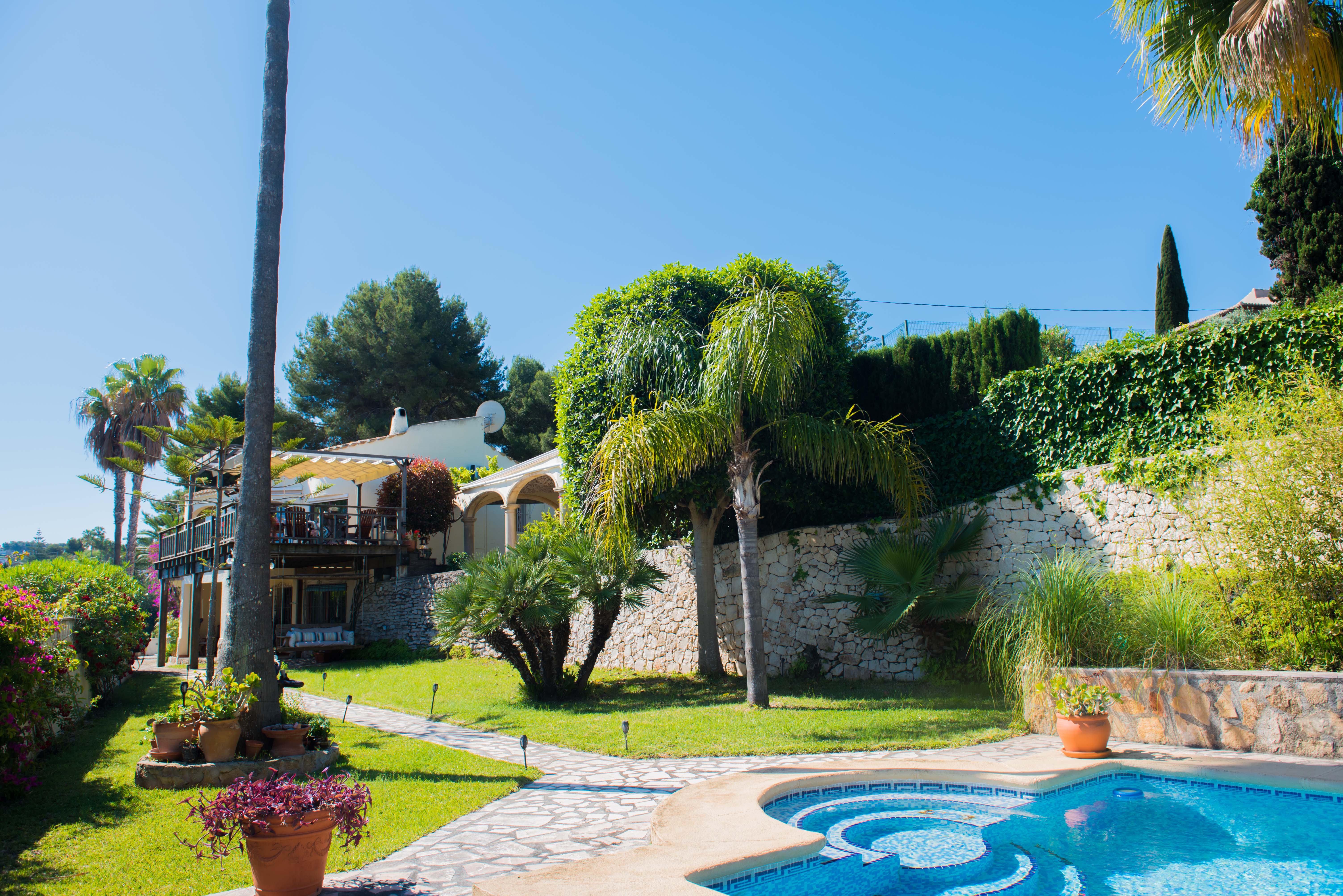 Vue sur le jardin avec piscine en mosaïque bleue et chemin de pierre menant à la villa de style méditerranéen.