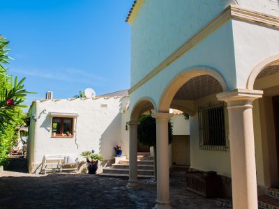 Vue extérieure de la villa avec des arcades en pierre et des murs blancs texturés, entourée de verdure méditerranéenne.