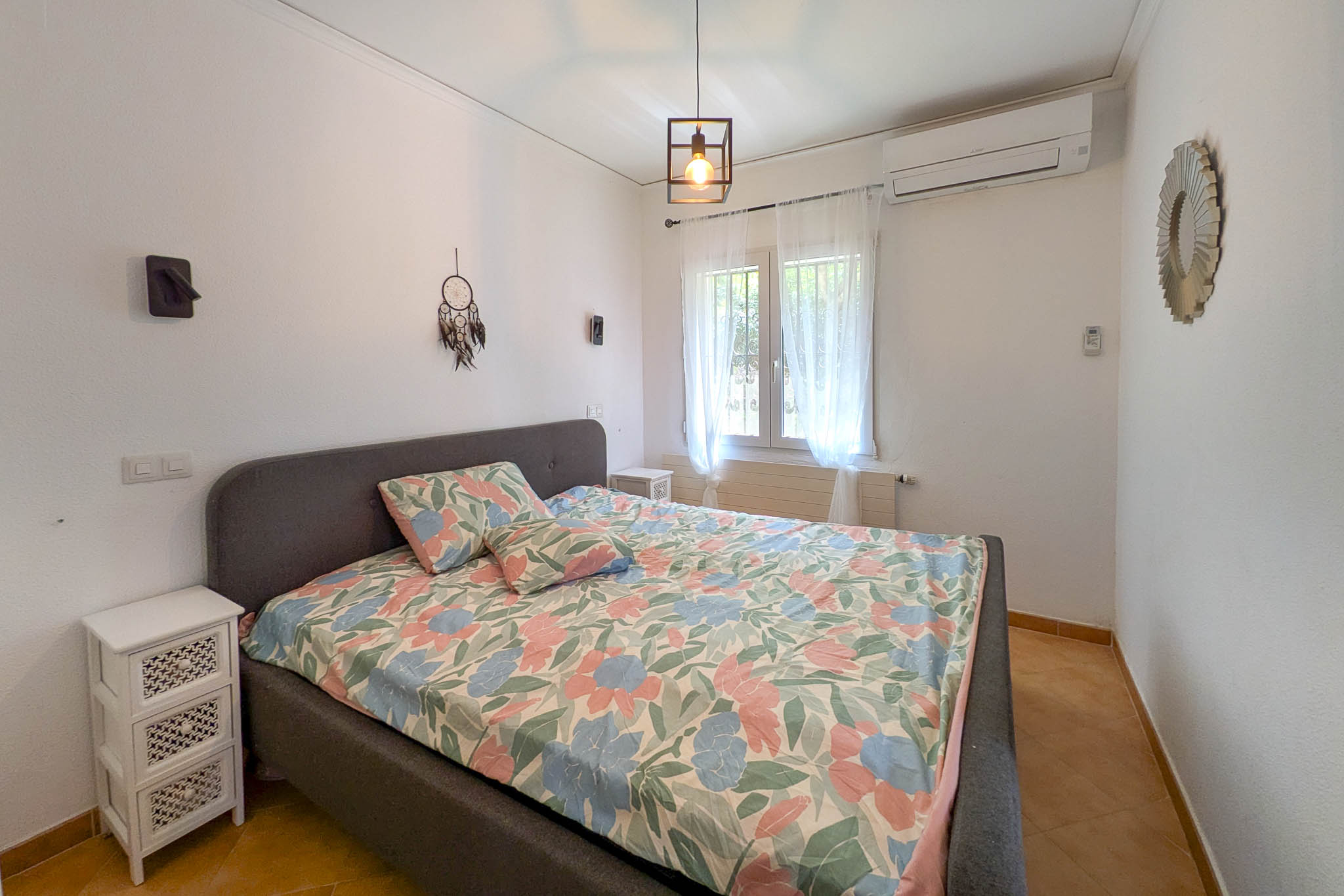 Chambre à coucher avec lit double, tête de lit rembourrée grise, climatisation split et sol en grès. La pièce dispose d'une fenêtre lumineuse et d'une suspension de style industriel.