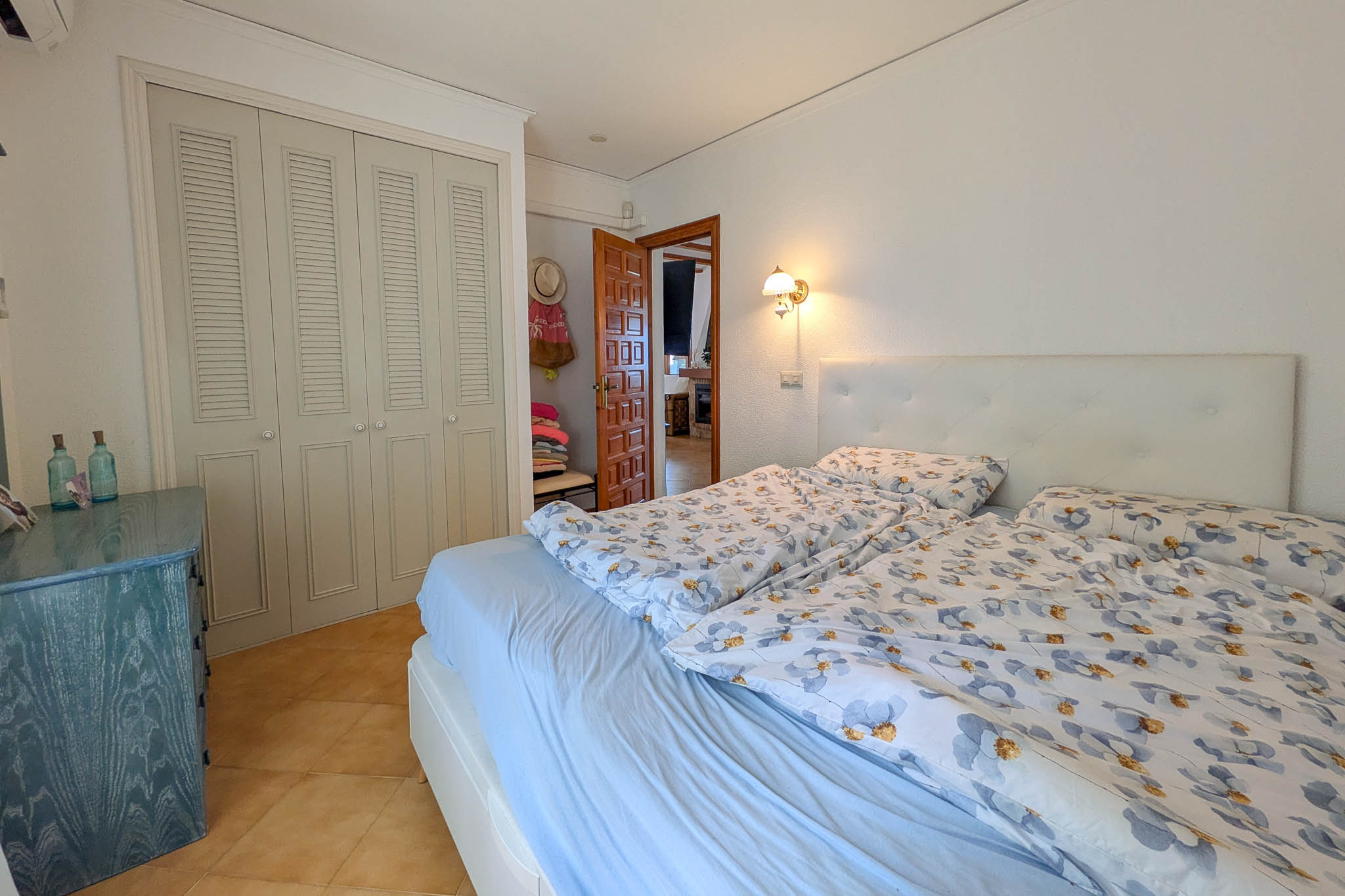 Chambre avec placards intégrés à persiennes, sol en grès cérame et tête de lit capitonnée. Une commode en bois bleu apporte une touche de couleur aux murs blancs.