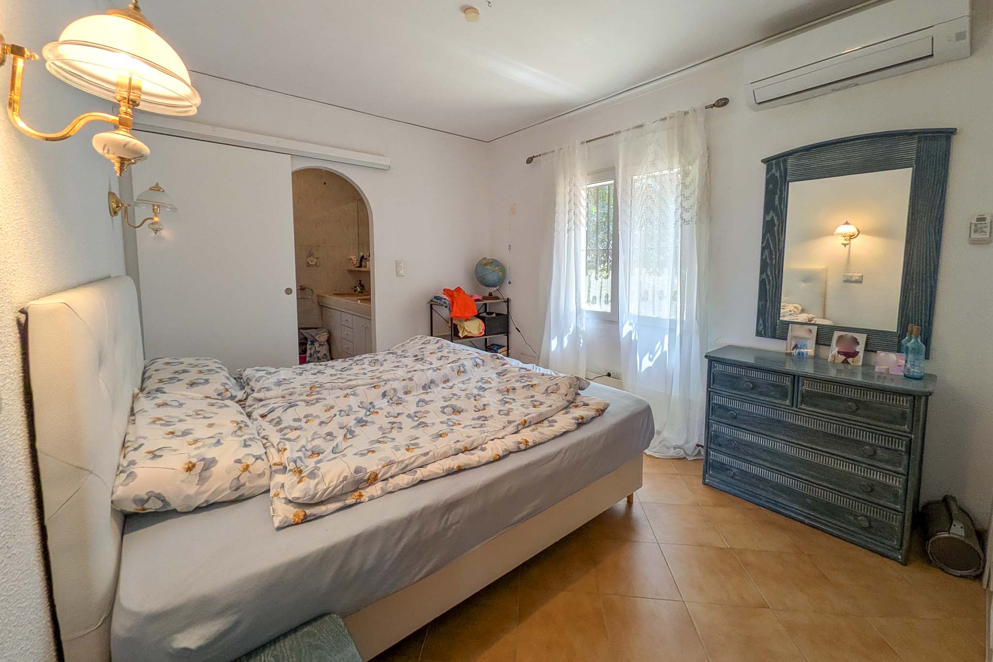 Chambre avec salle de bain attenante, climatisation split, sol en terre cuite et mobilier en bois. La pièce bénéficie d'une lumière naturelle constante.