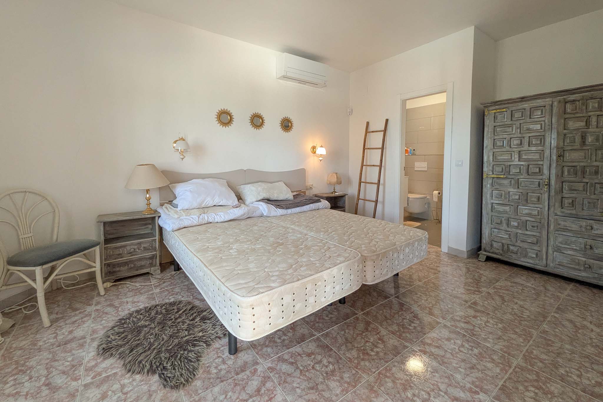 Chambre avec salle de bain attenante, sol en grès effet marbre, armoire rustique en bois sculpté et unité de climatisation murale.